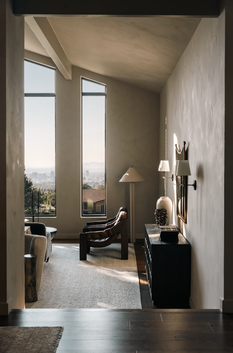 Living room with large vertical windows, leather armchairs, a floor lamp, side table, and a textured wall with lamps, overlooking a city skyline.