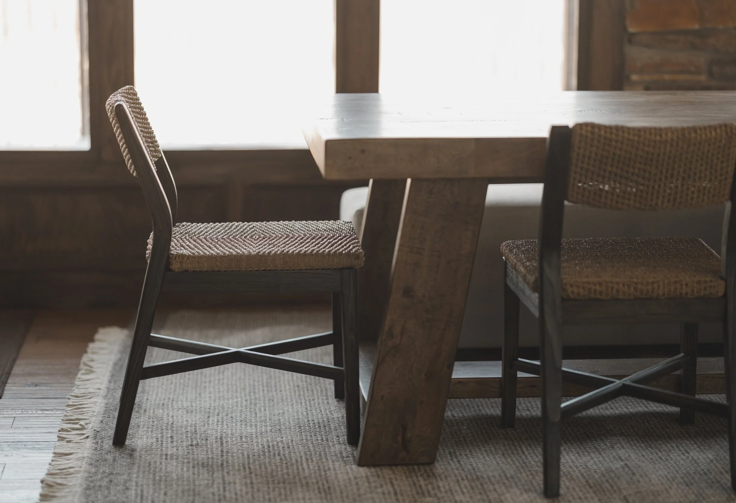 Interior of a rustic dining area with a wooden table and three woven chairs, natural light from windows, and a textured rug on wooden floors.