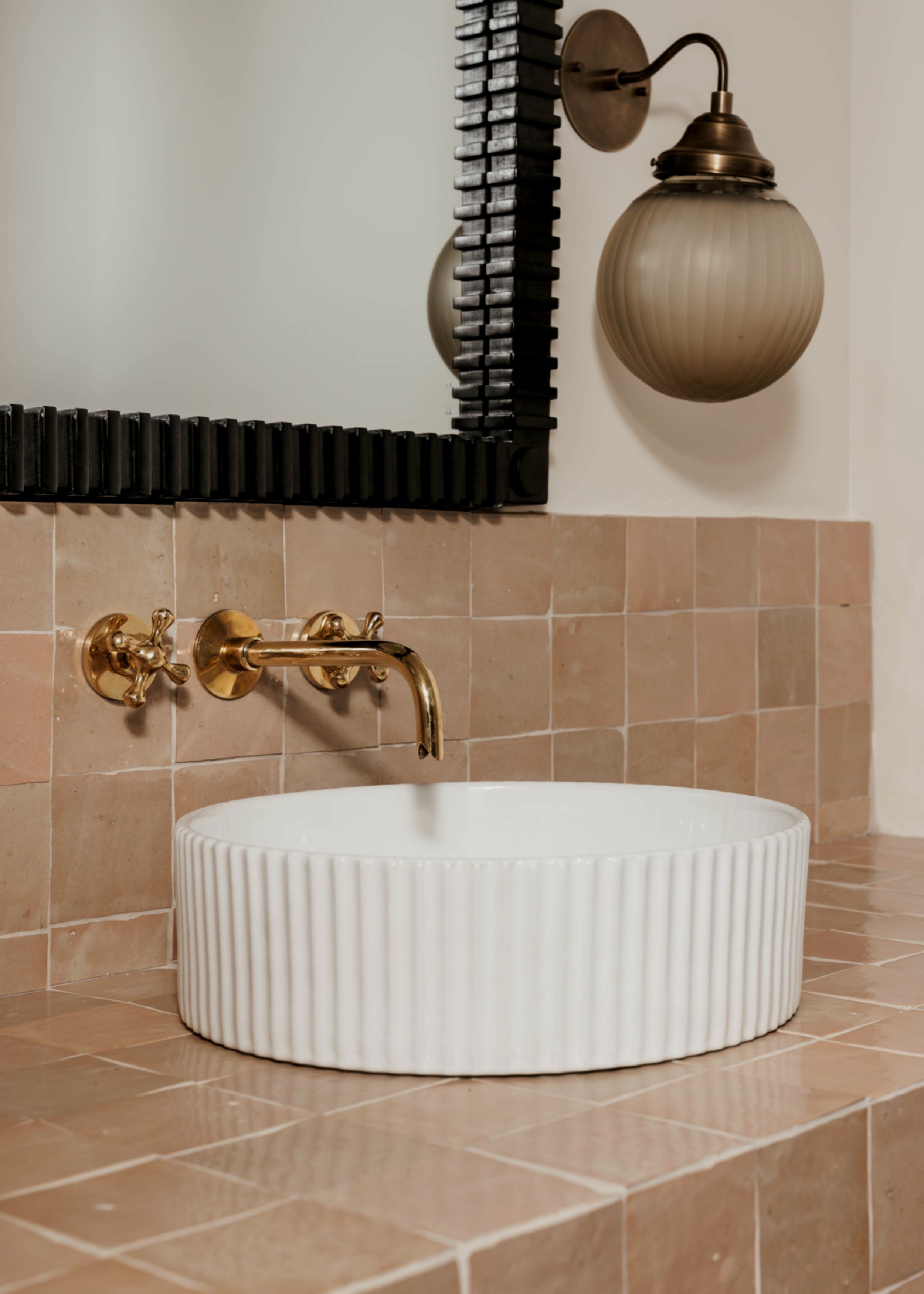 A bathroom sink with a white, ridged ceramic basin, a gold wall-mounted faucet, a large rectangular mirror with a black frame, and a vintage style wall sconce with a frosted glass shade.