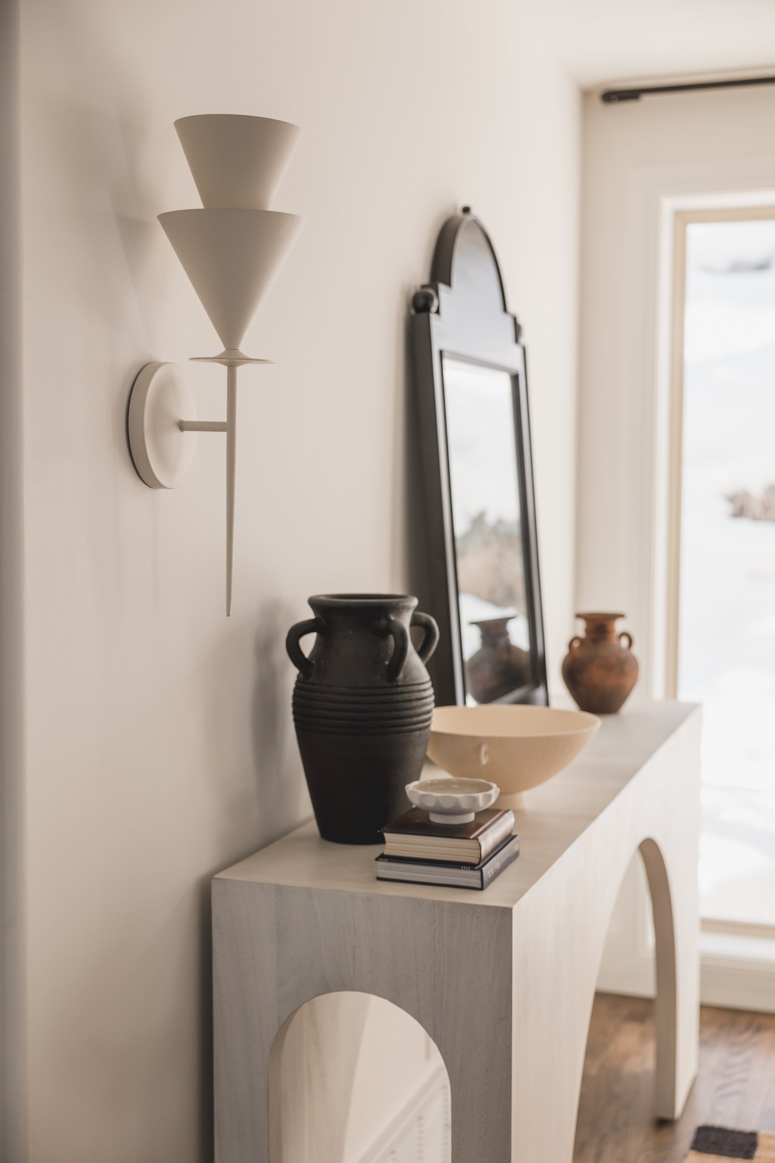 Photo of a minimalist interior with a white wall, a modern wall sconce, a beige sideboard, a large rectangle mirror, and decorative items including a black vase, a white bowl, a small white dish, and two brown vases.