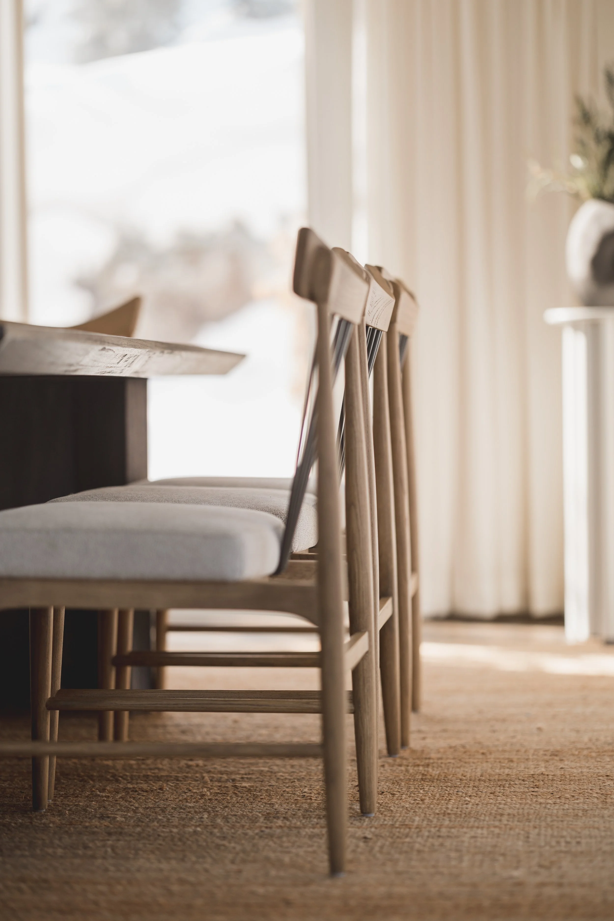 A row of wooden chairs with cushioned seats placed beside a wooden table, with sunlight streaming through a window with beige curtains.
