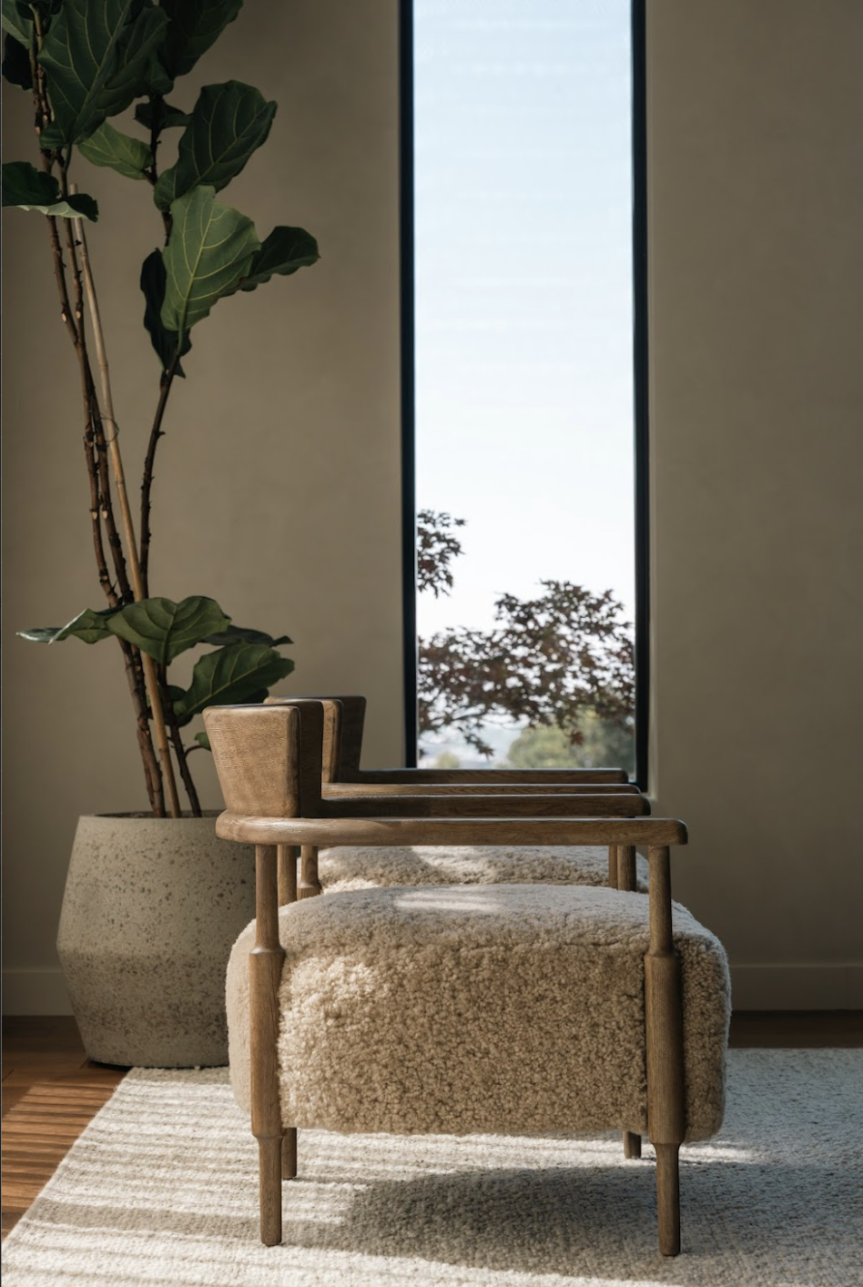 A cozy chair with a fluffy beige cushion in a bright room next to a tall potted plant and a large window showing trees outside.