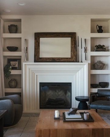 Living room with a fireplace, built-in shelves on both sides filled with black and decorative items, a large empty picture frame above the mantel, two chairs, and a wooden coffee table in front.