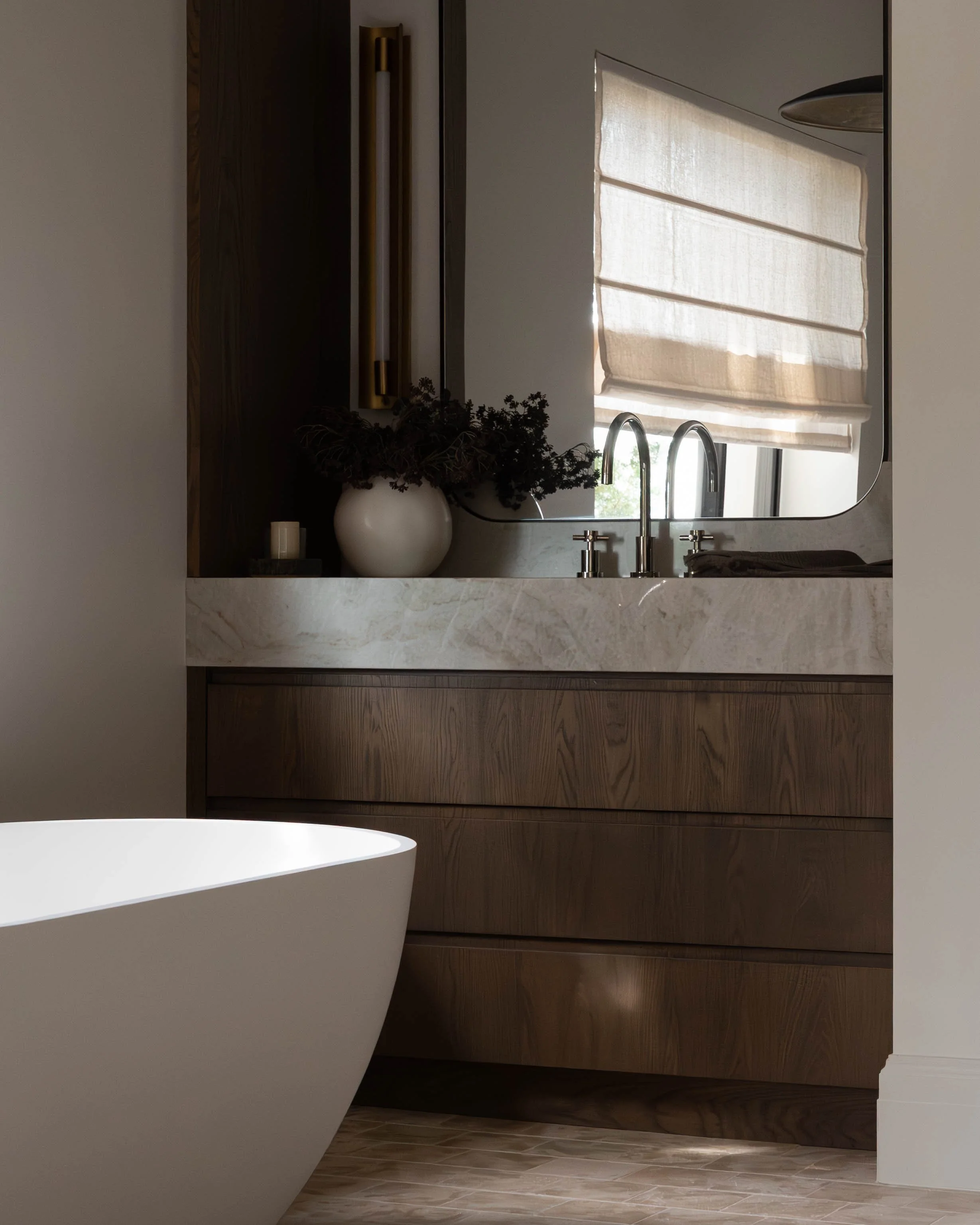 Modern bathroom with a white freestanding bathtub, a wooden vanity with drawers, a marble countertop, a large mirror, a window with shades, a silver faucet, and a white vase with dark foliage.