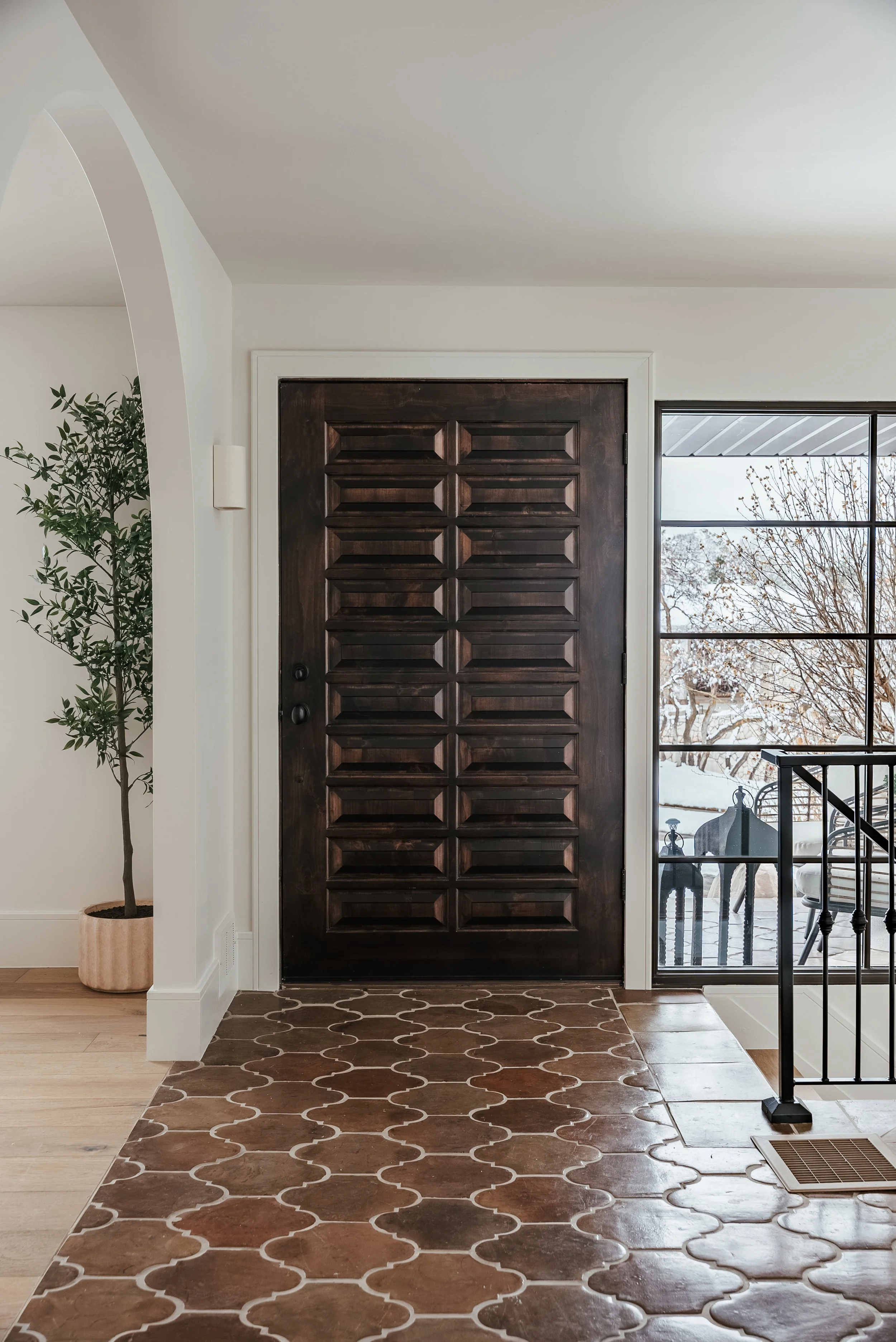 Indoor entryway with a dark wooden door, a potted plant on the left, a large window with a balcony and outdoor scenery on the right, and patterned tile flooring.