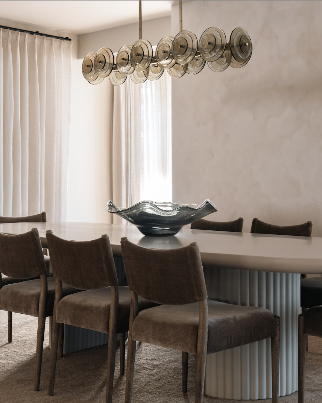 Modern dining room with a white oval table, brown upholstered chairs, a glass bowl centerpiece, and a unique ceiling light fixture with multiple round shades.