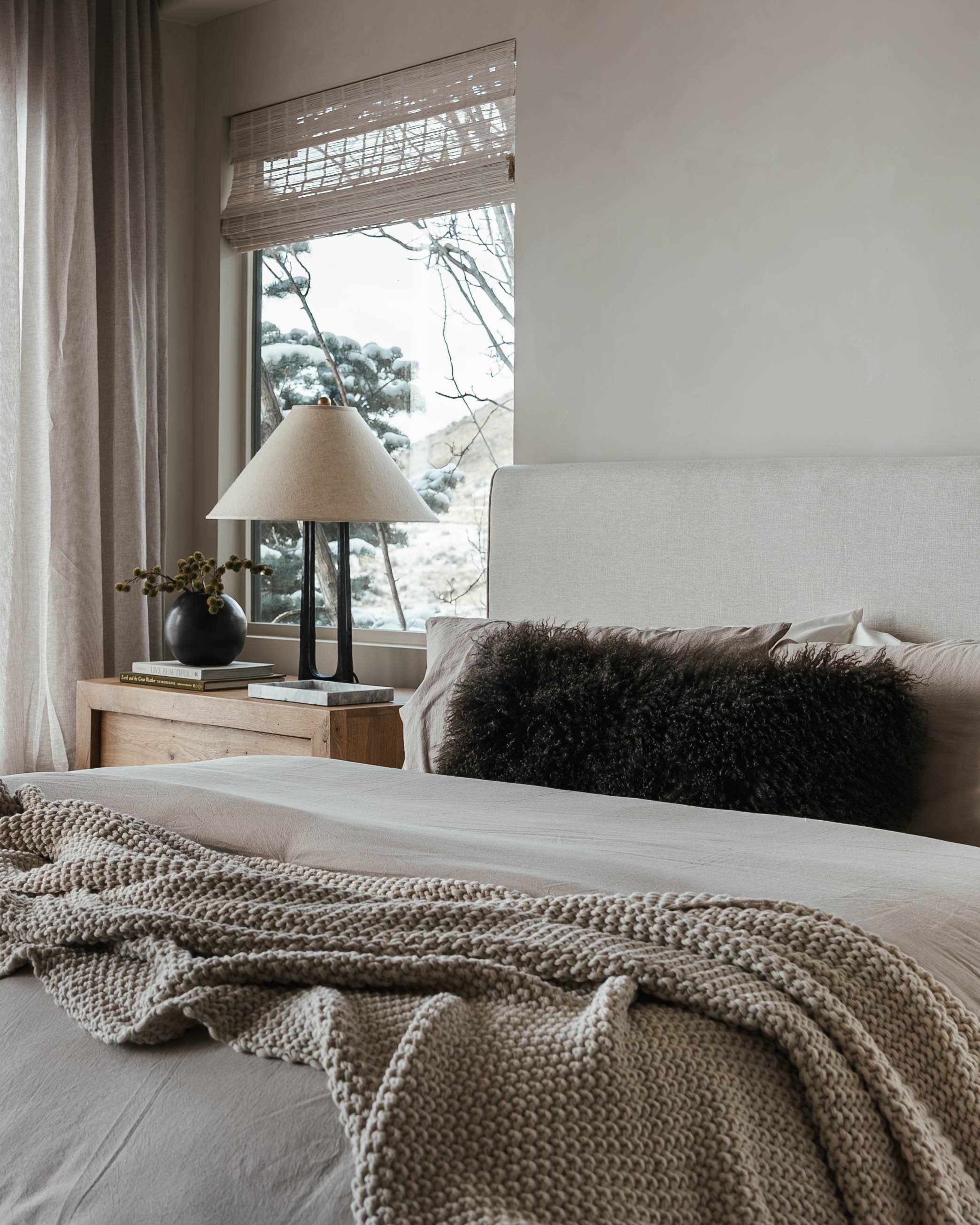 A cozy bedroom with a bed covered in neutral-colored linens, a textured throw blanket, and a black furry pillow. There is a wooden nightstand with a black vase with greenery, and a modern table lamp. A large window shows a winter scene with snow-cove