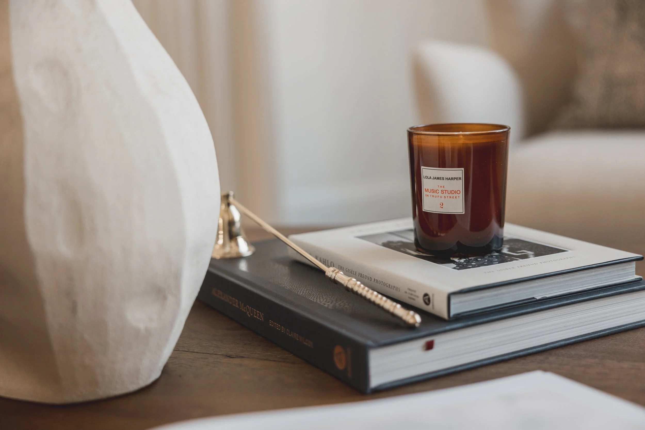 Close-up of a table with a large white ceramic vase, a stack of two books, a brown candle holder with a brown candle on top, and a pen.