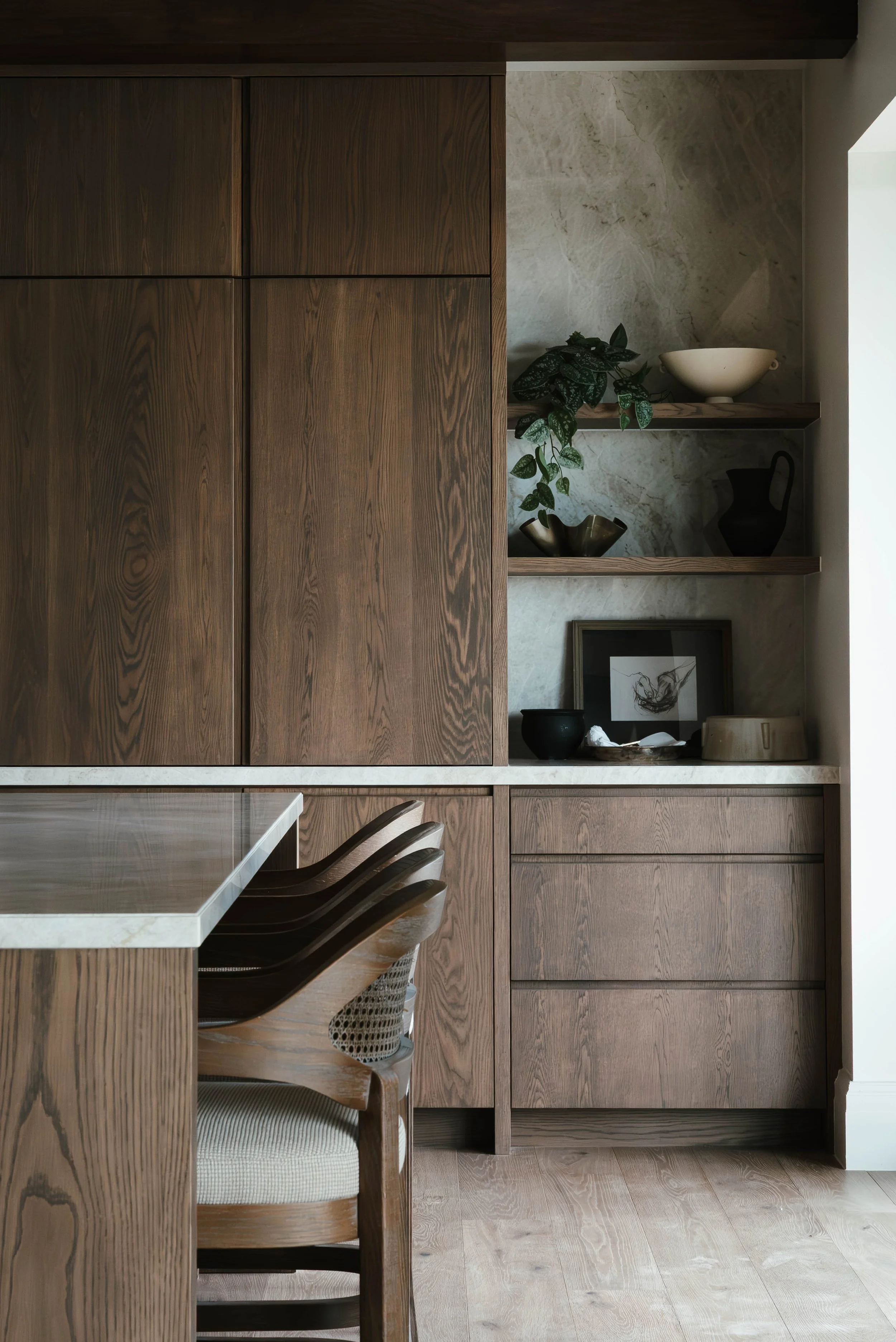 A modern kitchen with dark wood cabinetry, marble countertops, and open shelves displaying decorative items, a framed picture, and a potted plant.