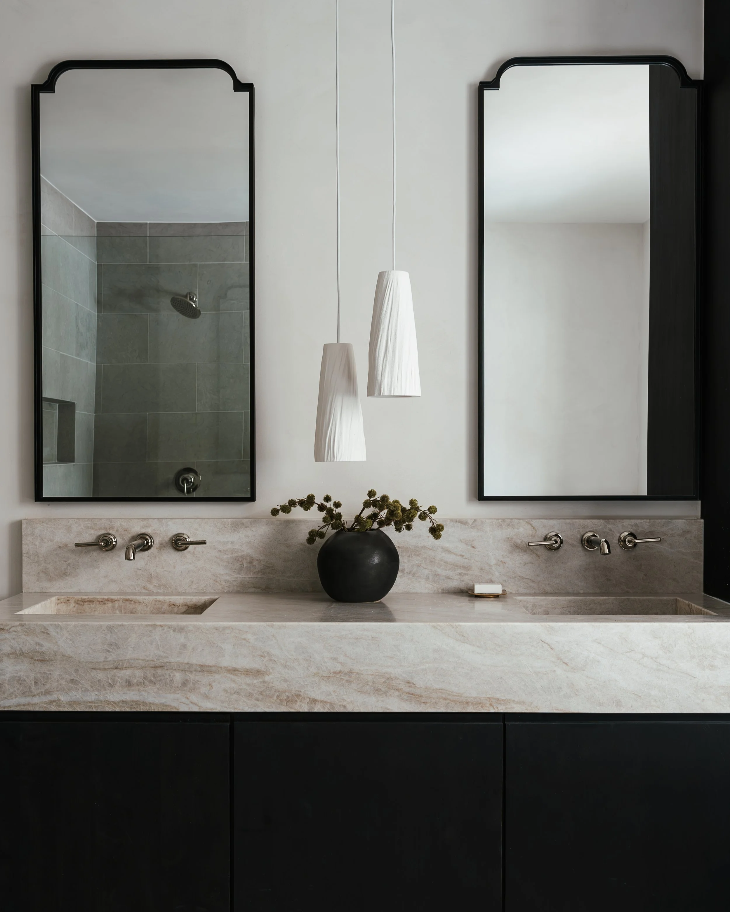 Modern bathroom vanity with two large mirrors, hanging pendant lights, a black vase with green foliage, and a beige marble countertop with two sinks.