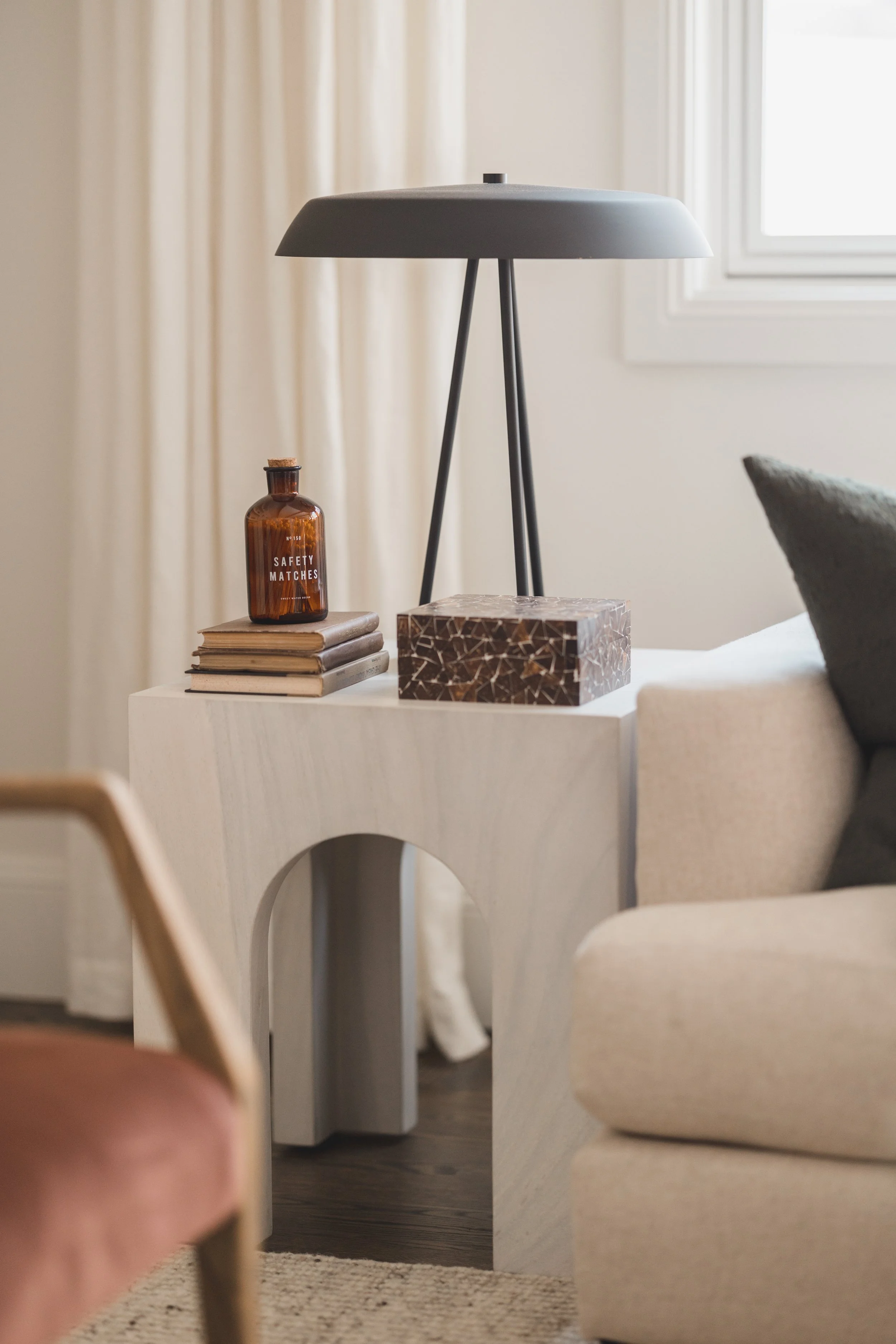 A modern living room side table with a wooden design. On top are four stacked books, a brown bottle labeled 'Safety Matches,' and a decorative rectangular box. There is a tall, dark-gray lamp with a tripod base on the table. Part of a beige sofa and 
