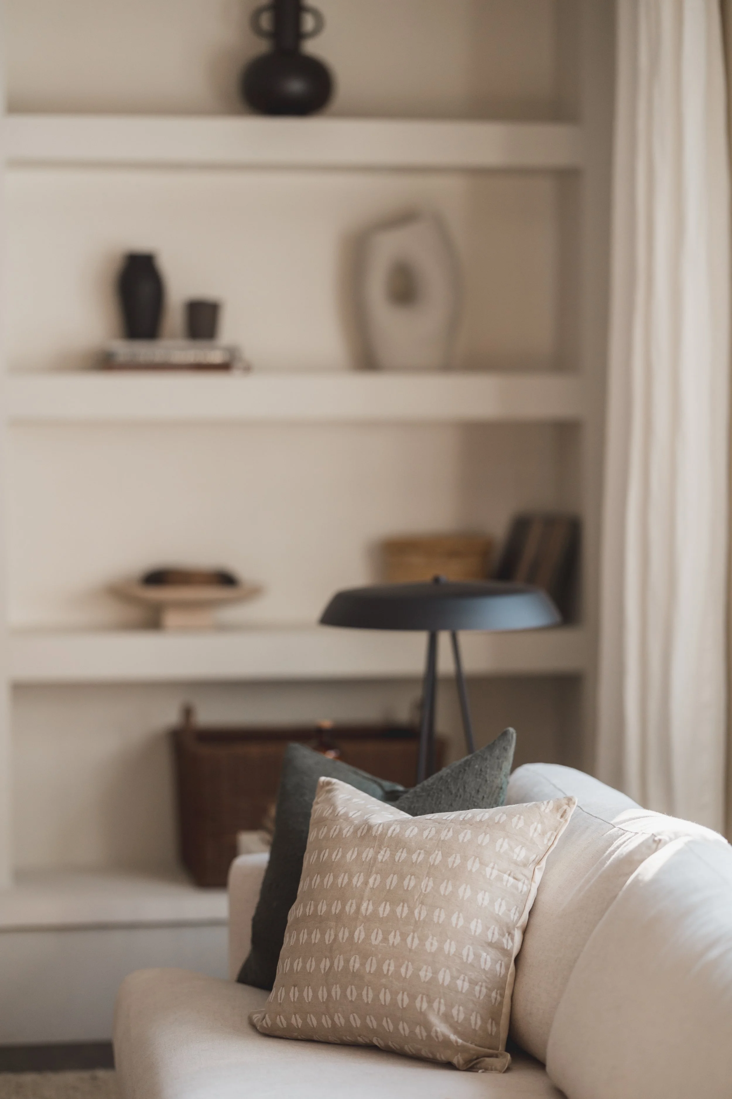 A living room with a white sofa and beige and dark green pillows, a black floor lamp, and built-in white shelves with decorative items and books. There are cream curtains on the right side.