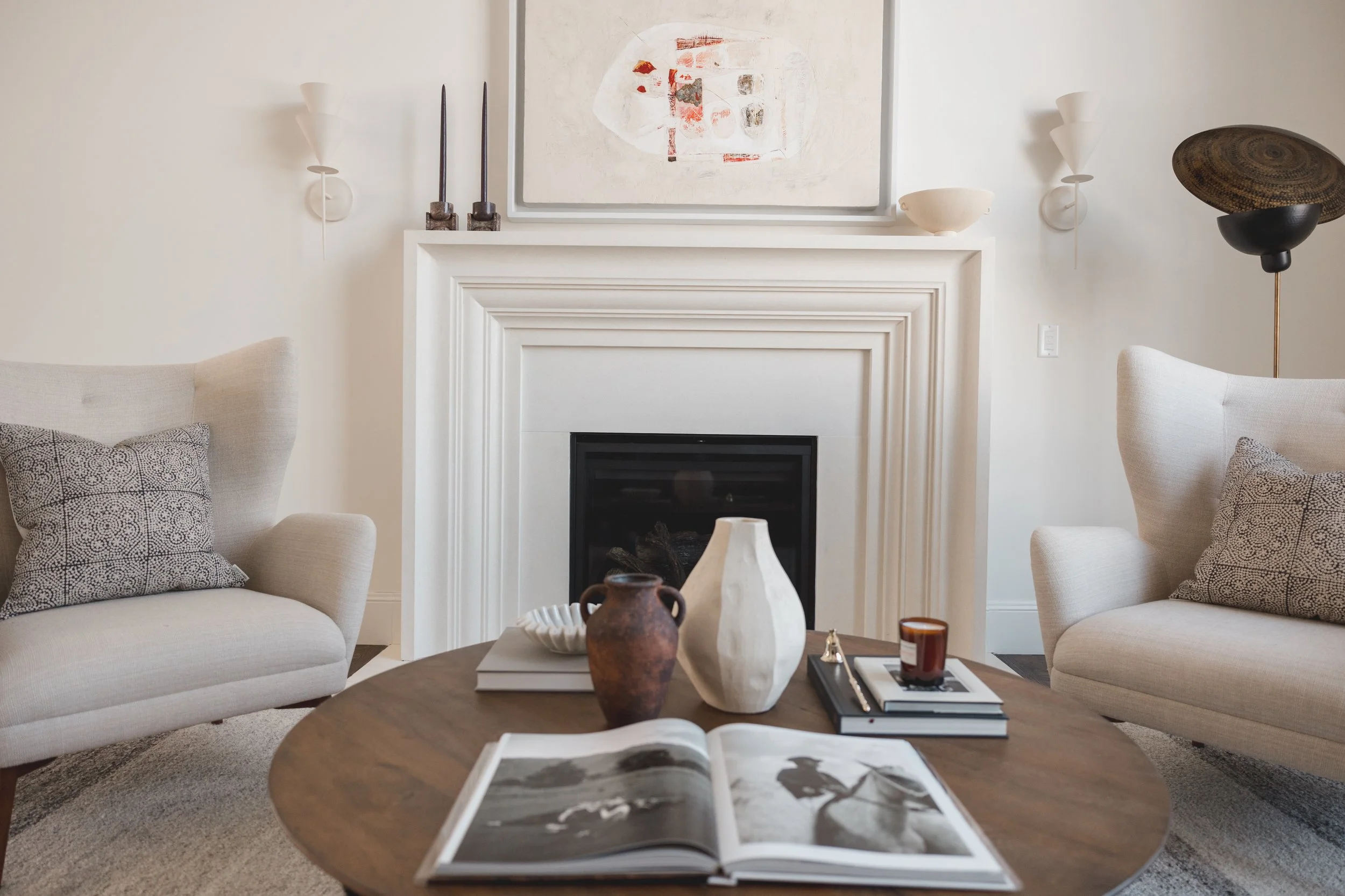 Living room with a fireplace, two armchairs, a round wooden coffee table with decorative vases, books, and a candle, and artwork above the fireplace.