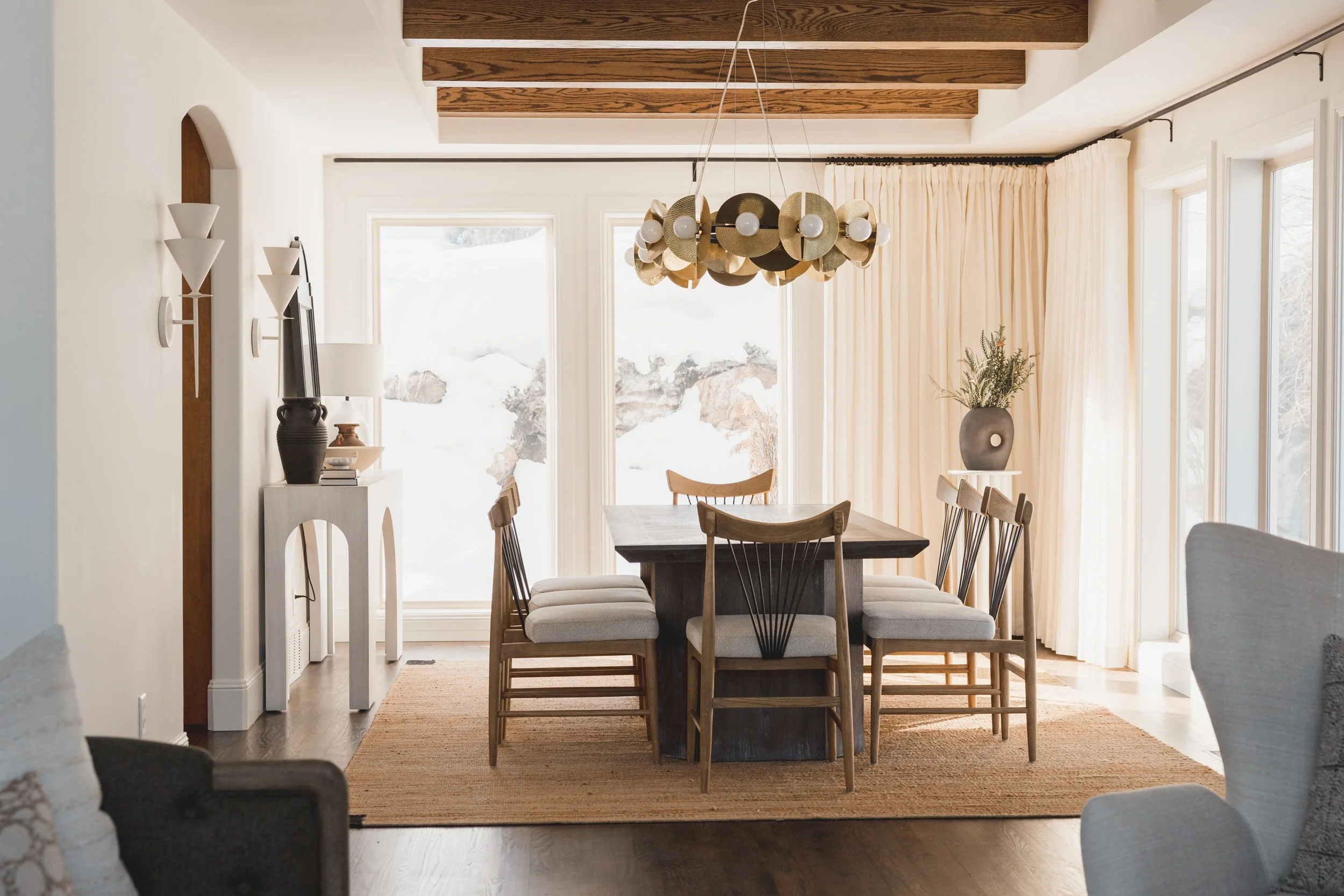 Dining room with a dark wood table, six wooden chairs with light cushions, a wooden beamed ceiling, a modern chandelier, large windows with cream curtains, a fireplace with decorative objects, and a large vase with greenery.