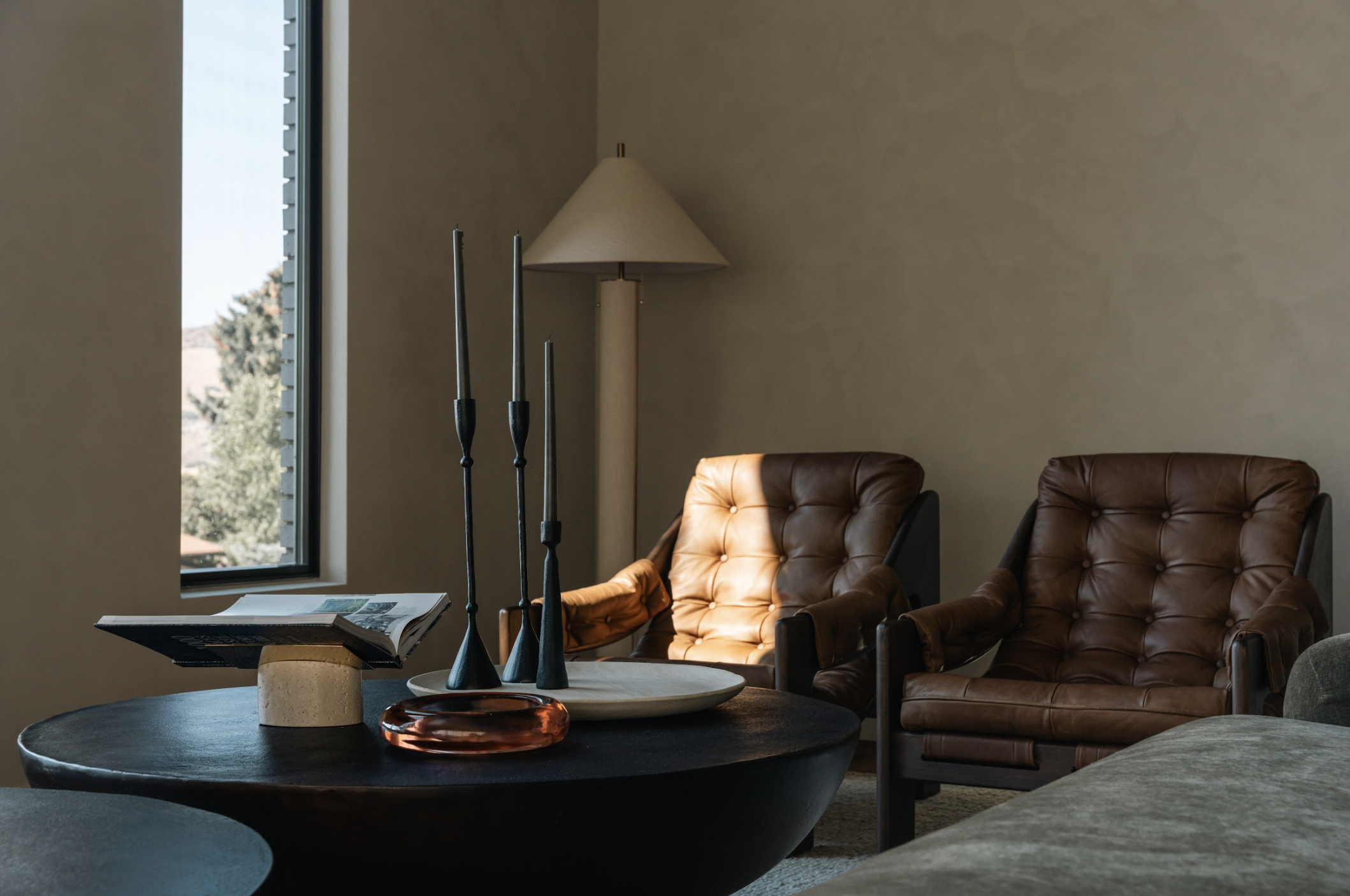 Living room with two brown leather armchairs, a window with outside trees, a white floor lamp, and a black coffee table with decorative items and an open magazine.
