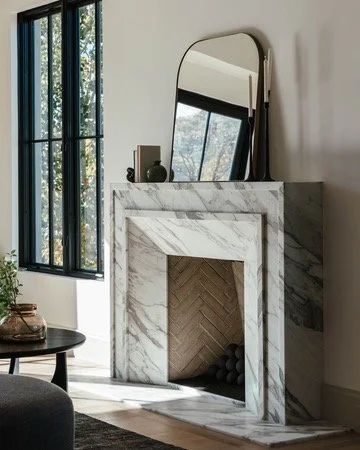 Modern fireplace with a marble mantel, a large mirror resting against the wall, a window with black metal frames, a small table and decorative objects nearby.