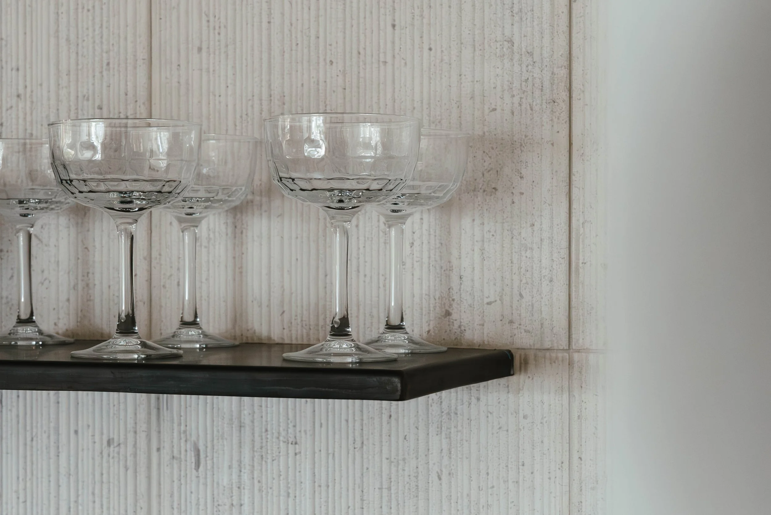 Multiple empty cocktail glasses with stems on a black shelf against a textured white wall.