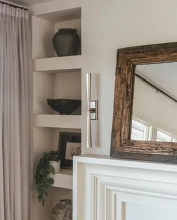 Interior corner of a room with built-in white shelves, decorative items, a framed picture, green plant, and a mirror with a wooden frame.