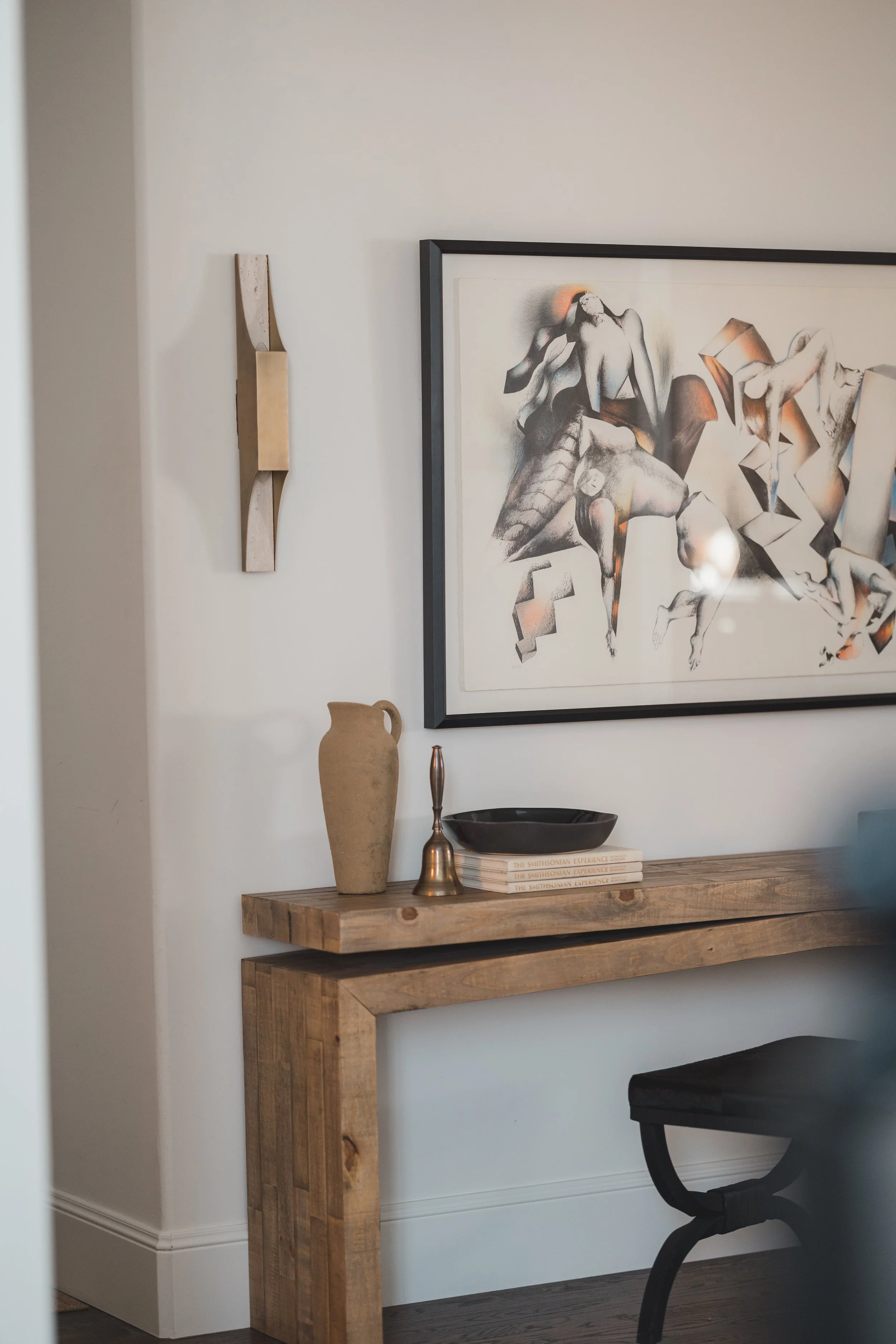 Interior of a room with a wooden console table, a large framed abstract art piece, decorative vases, a bell-shaped sculpture, and a stack of books on the table.