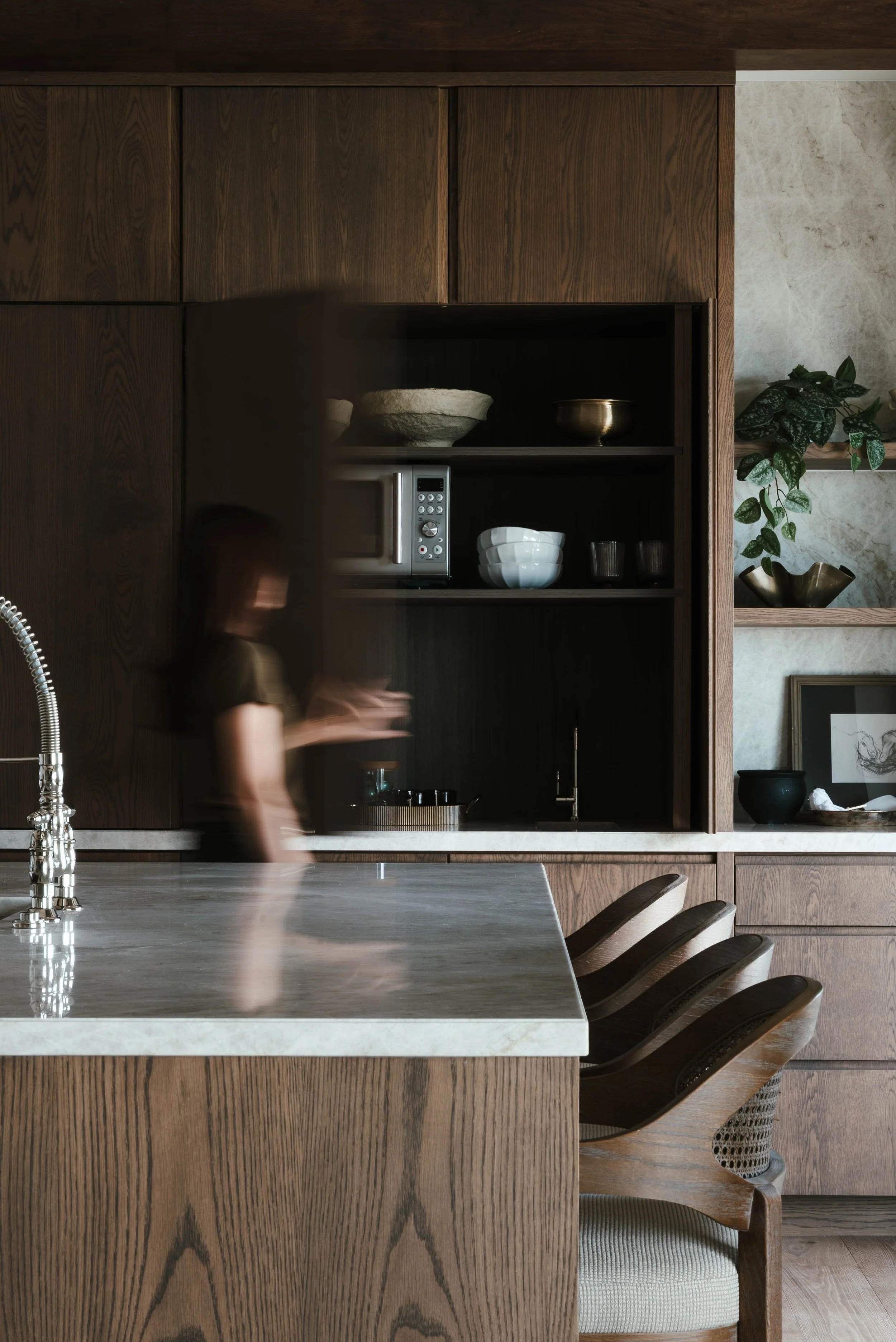 A modern kitchen with dark wood cabinetry, a white marble countertop, and a person blurred in motion standing near the counter.
