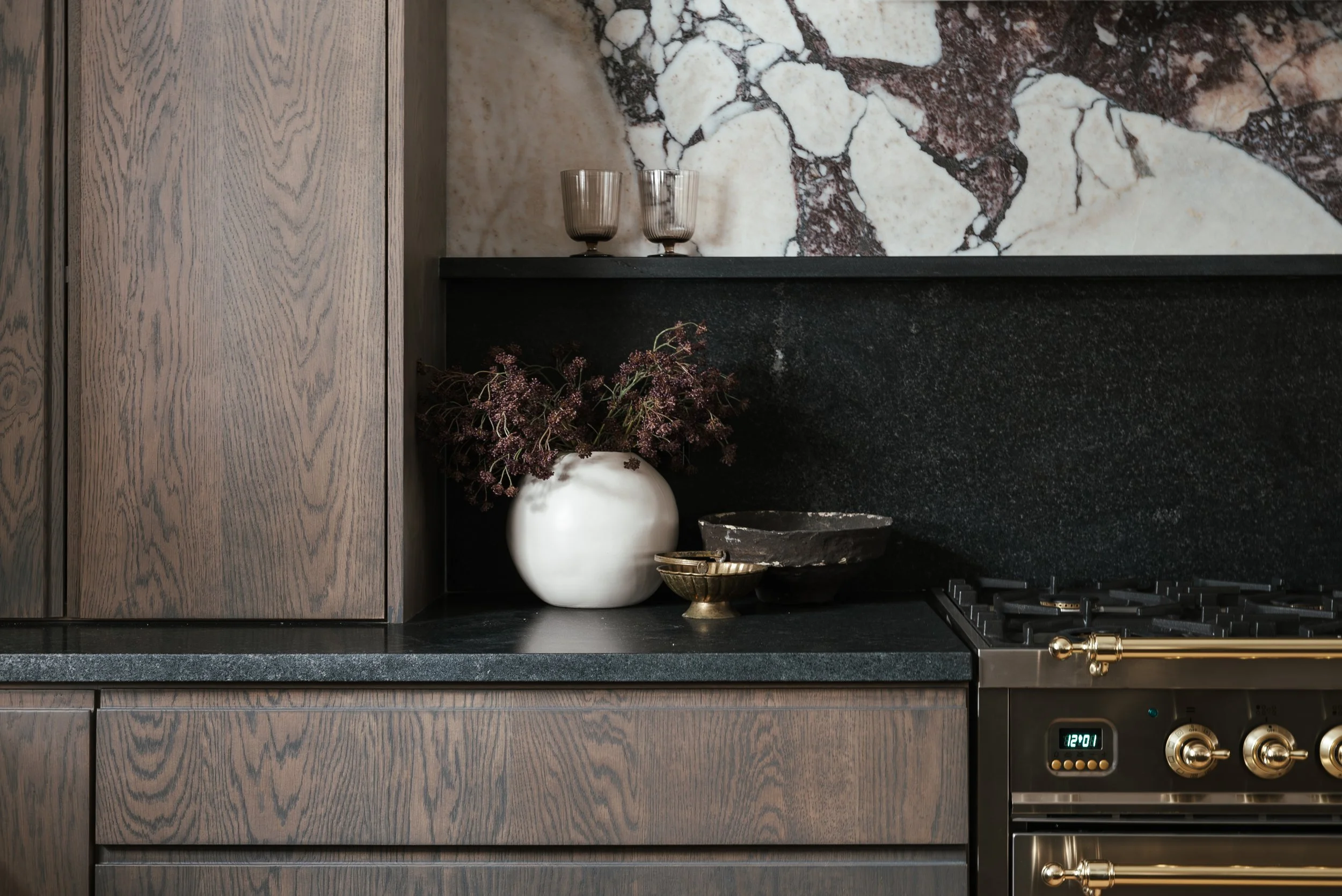 Close-up of a modern kitchen countertop with a white vase and purple flowers, black stone bowls, and a stove with gold accents against wooden cabinets and a marble backsplash.