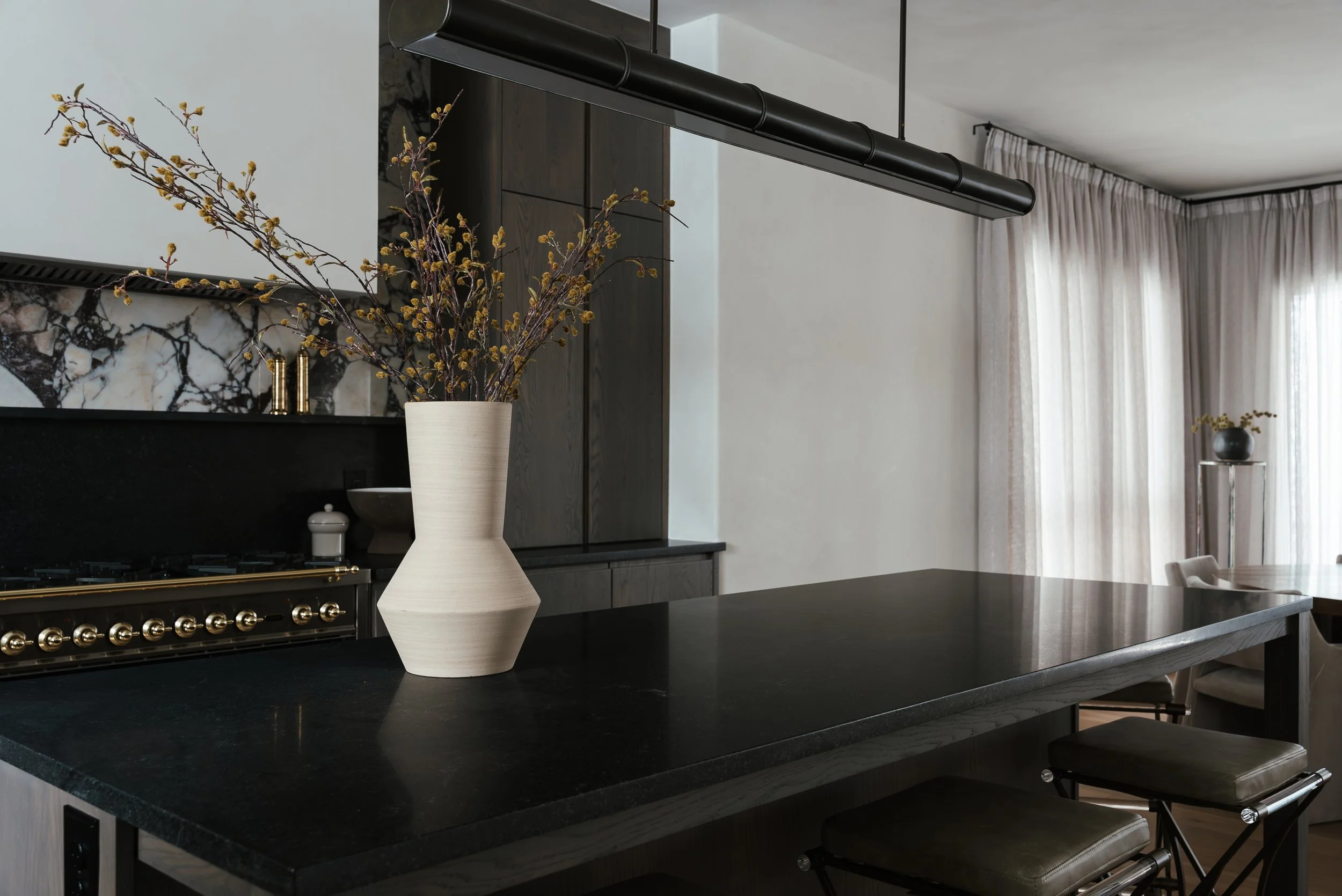 A modern kitchen with a black countertop, a large white vase with purple and yellow flowers, dark cabinets, beige curtains, and a dining area with chairs in the background.