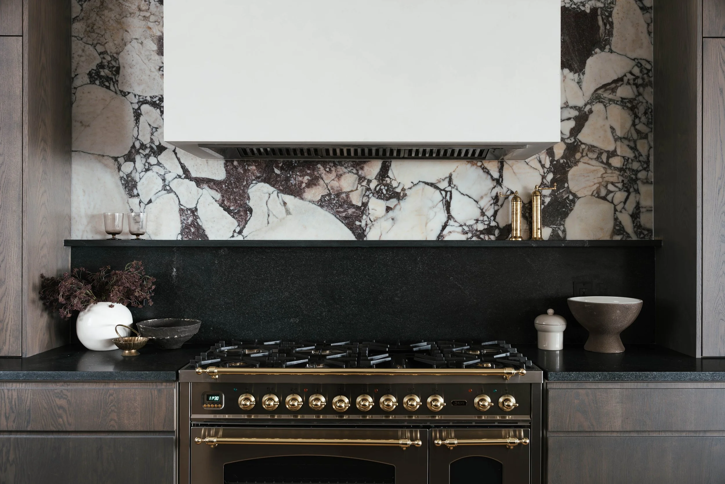 Modern kitchen with black marble countertops, stainless steel stove with gold knobs, marble backsplash, white cabinetry, and decorative items including glasses, bowls, and small containers.