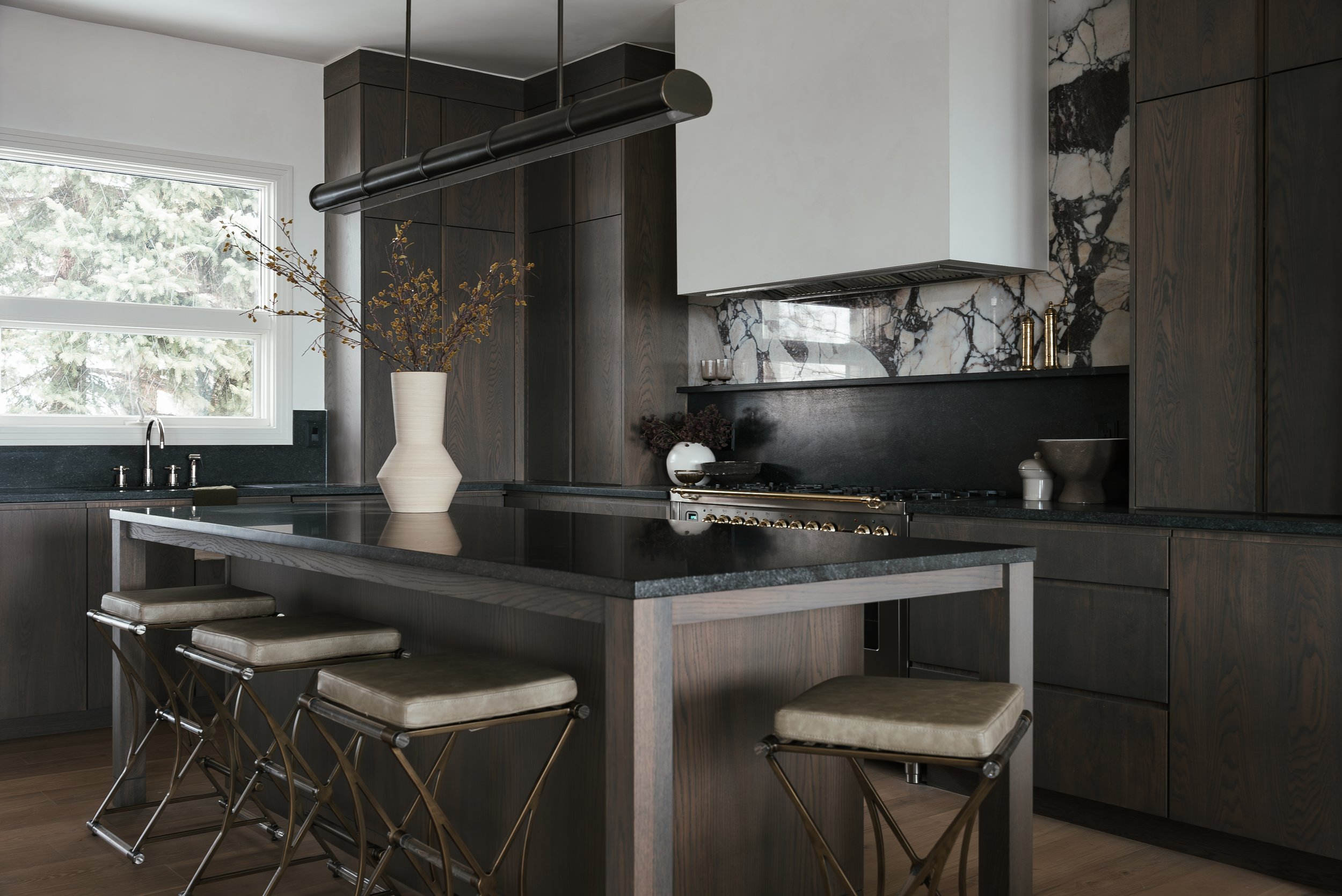 Modern kitchen with dark wood cabinets, black countertop, marble backsplash, and an island with beige cushioned stools, marble vase with branches, window showing greenery outside, and minimal decorative items.