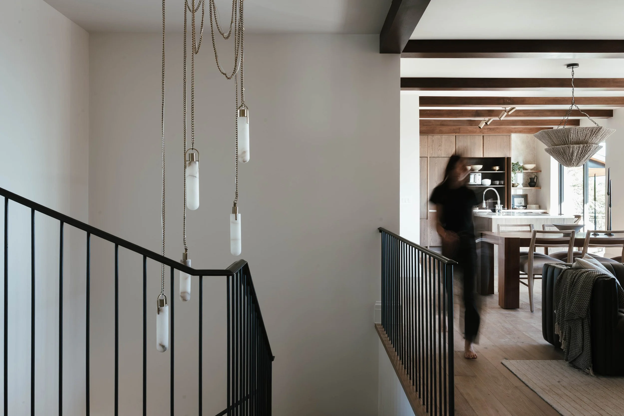 Interior of a modern home with a staircase, hanging pendant lights, and an open kitchen and dining area in the background, with a blurred person walking through.