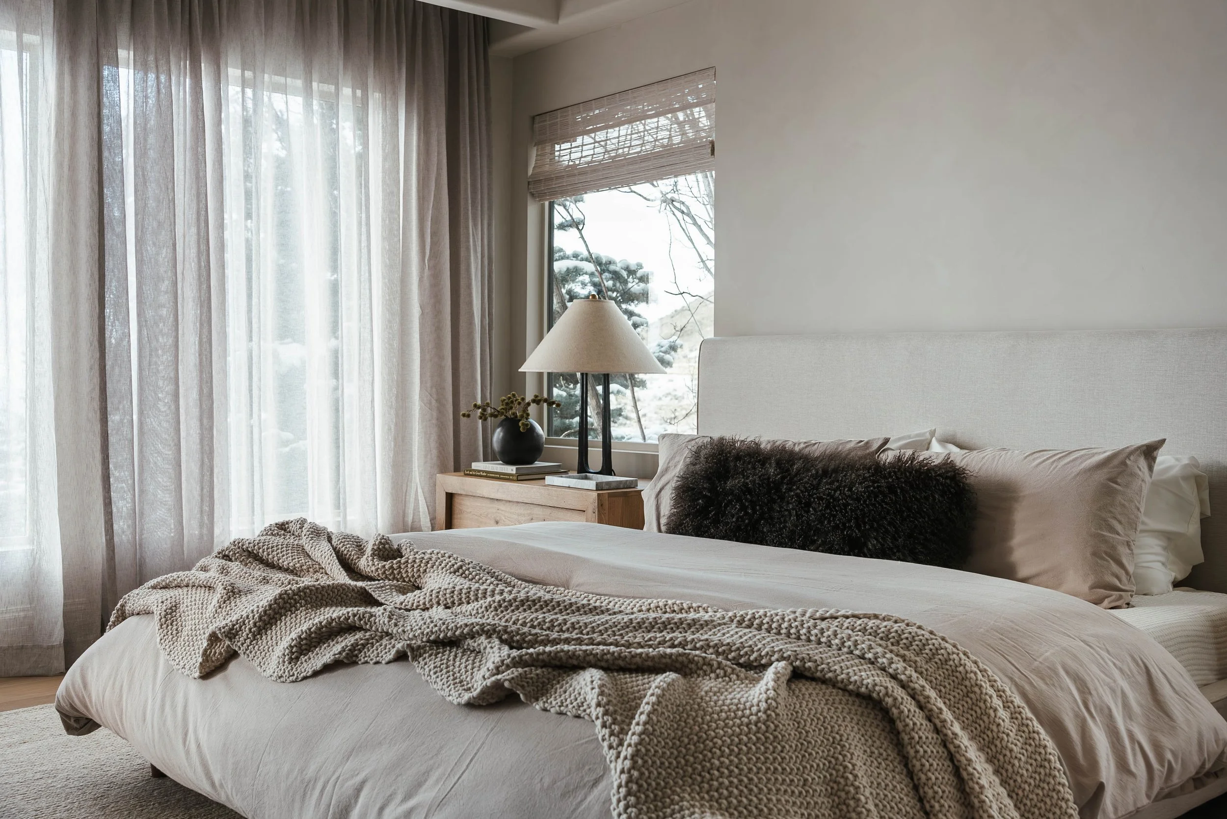 A cozy bedroom with a neatly made bed, beige and cream bedding, a textured throw blanket, pillows including a dark furry one, a wooden nightstand with a lamp, a black vase with greenery, and a window showing snow-covered trees outside.