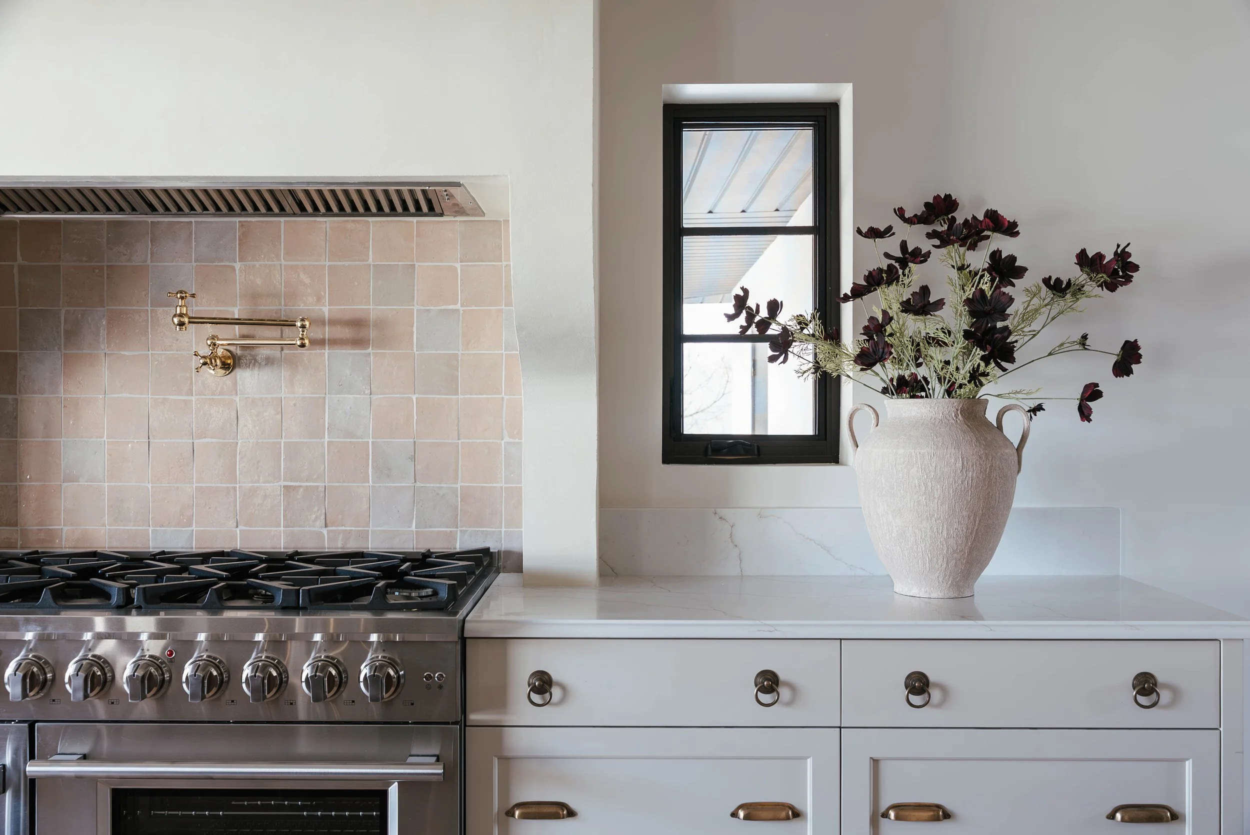 Kitchen with beige tiled backsplash, stainless steel oven and stove, white countertop, black framed window, and large white vase with dark purple flowers