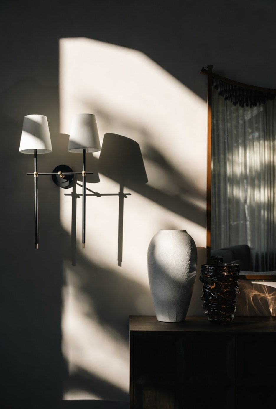 Interior corner with a wall-mounted lamp casting shadows, a large textured white vase, a dark glossy vase, a wooden cabinet, and a mirror reflecting curtains and sunlight.