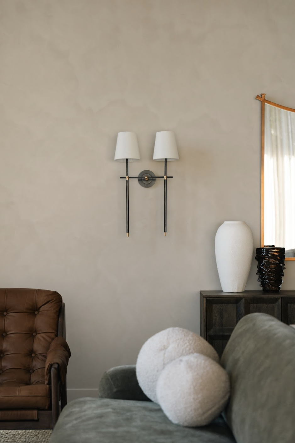 A living room corner with a brown leather armchair, a green velvet sofa with textured white pillows, a wooden side table with a white ceramic vase and a black decorative object, a large mirror with a wooden frame, and a wall-mounted two-shade sconce 