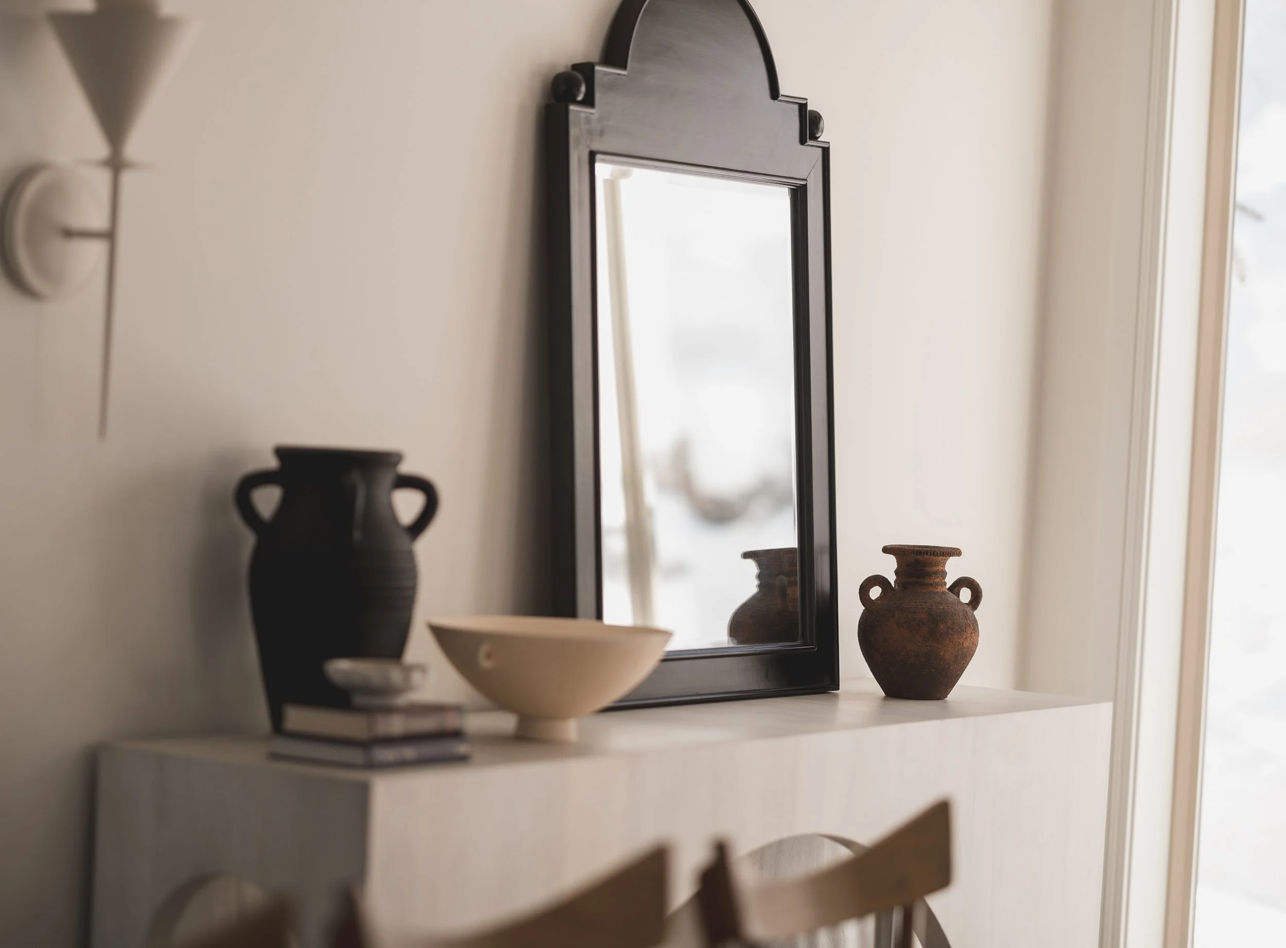 Interior view of a white bookshelf with decorative pottery vases, a bowl, and a framed mirror. Natural light coming through a nearby window.
