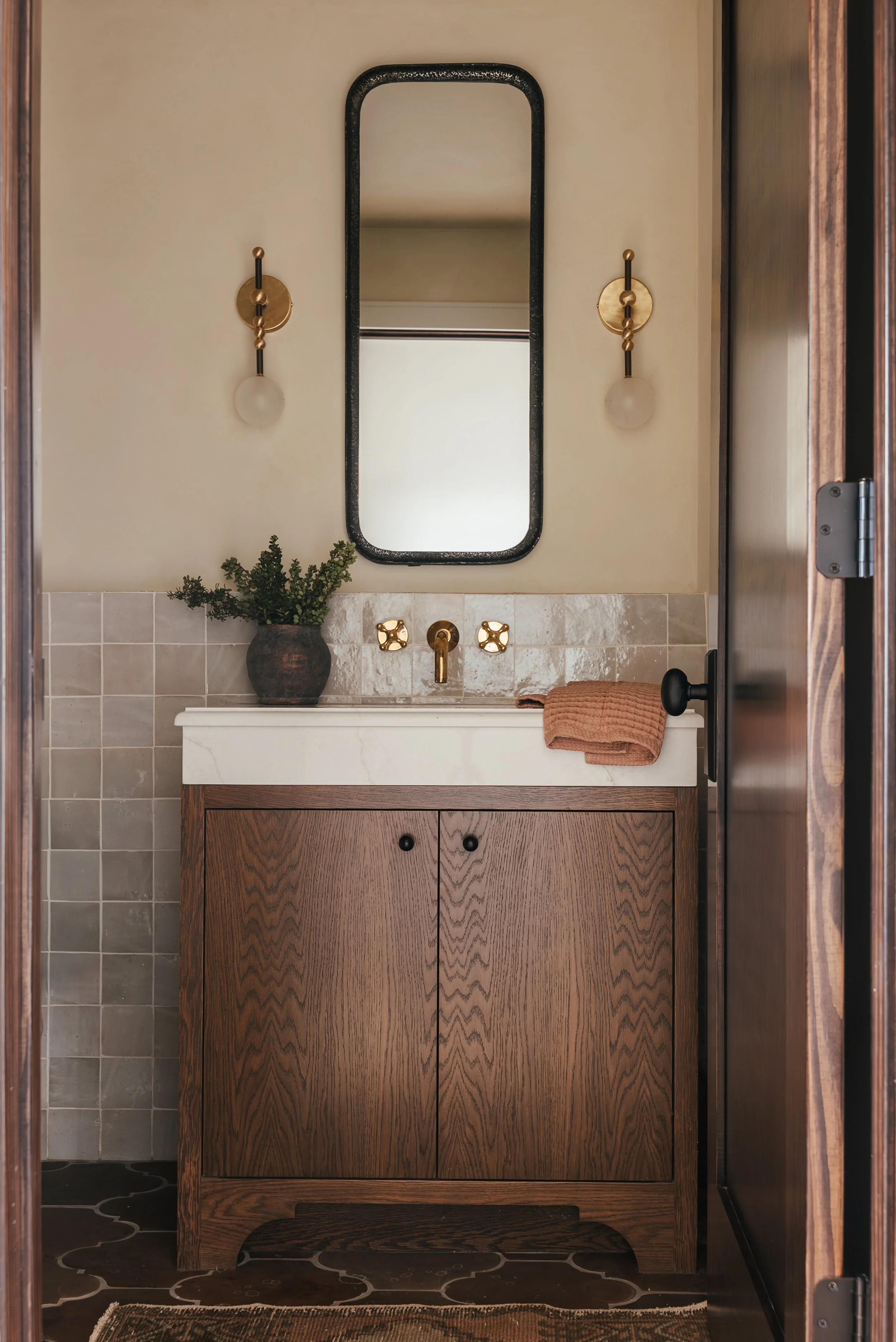 Bathroom with a wooden vanity, small sink, gold fixtures, a mirror, two wall sconces, a plant in a dark pot, and a towel on the counter.