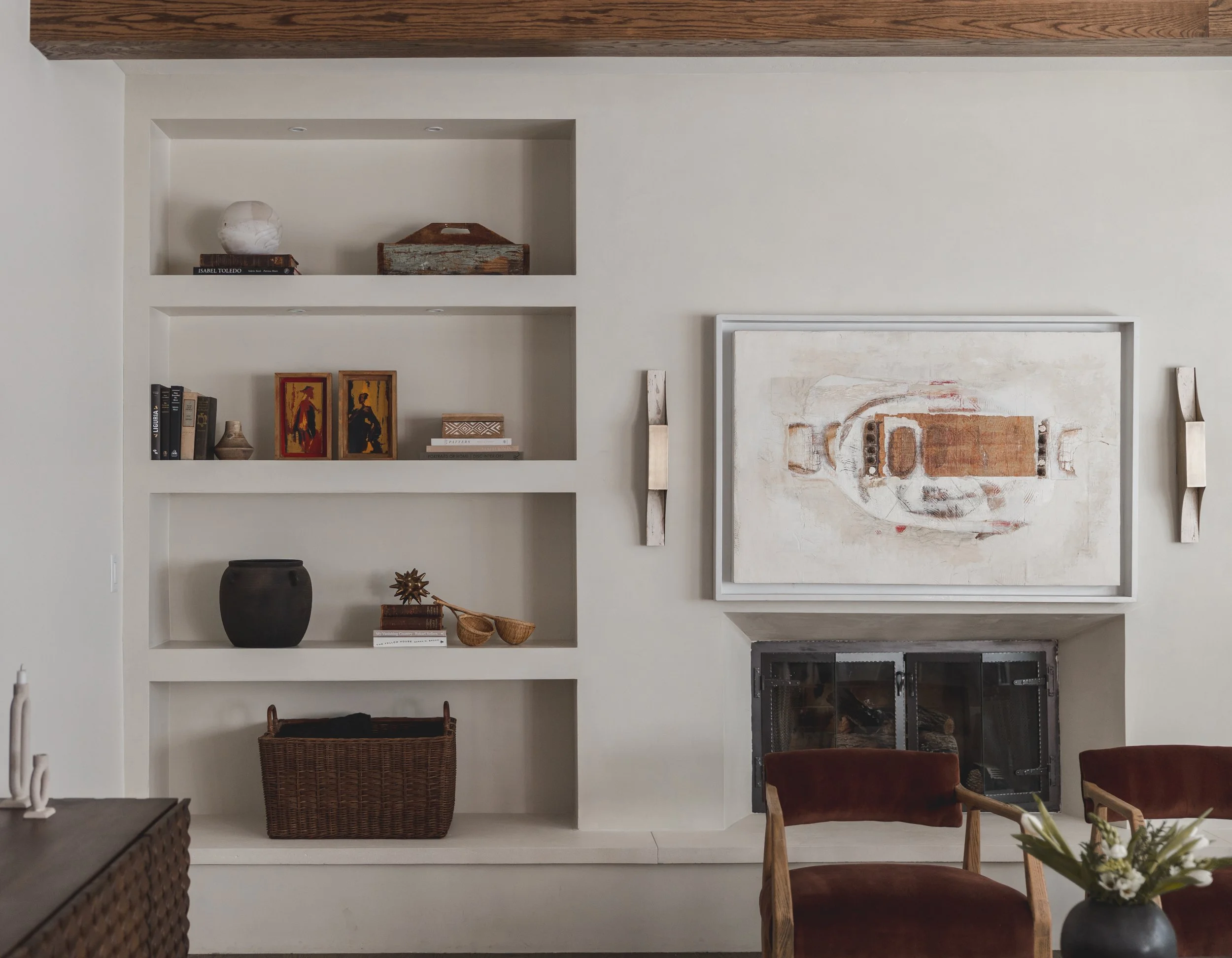 A close-up of a built-in wall shelf with decorative items, a large abstract painting hanging above a fireplace, and two upholstered chairs in a living room.