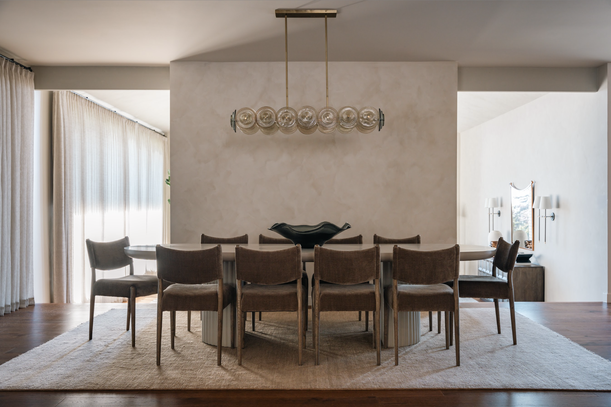 Modern dining room with a large round table surrounded by nine brown upholstered chairs, a black decorative bowl in the center, a light fixture hanging above, and sunlight filtering through sheer curtains on the left.