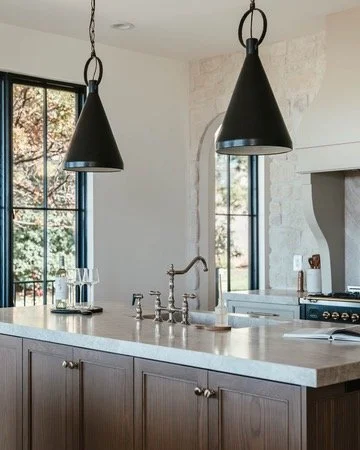Modern kitchen island with a white countertop, dark wood cabinets, and a stainless steel sink with a curved faucet. Two black pendant lights hang above. Large window and door with glass panels allow natural light, light-colored walls, and a stove wit