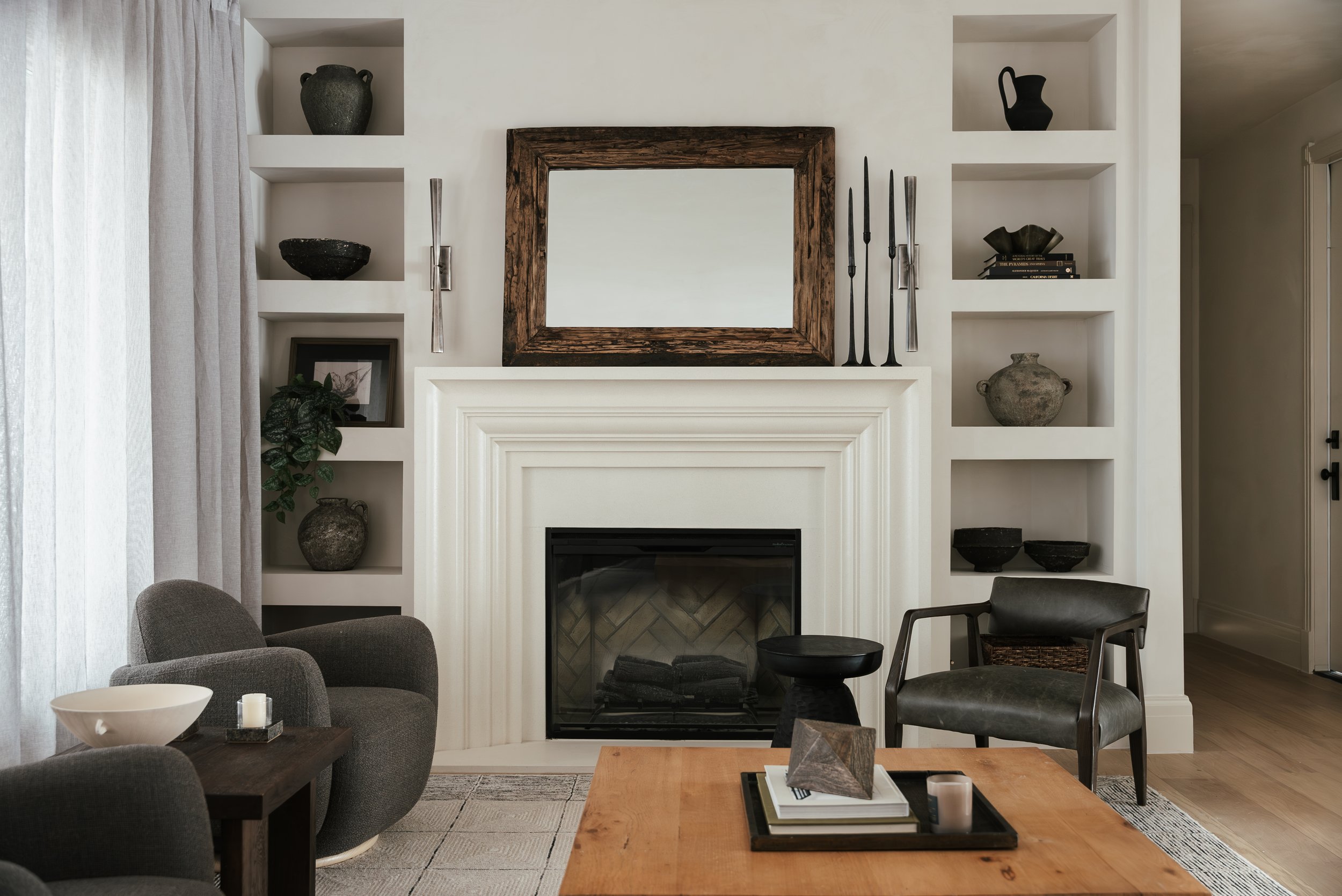 Living room with white fireplace, framed mirror, black and gray decor, built-in shelves with sculptures and plants, gray armchair, wooden table with decor, and light curtains.