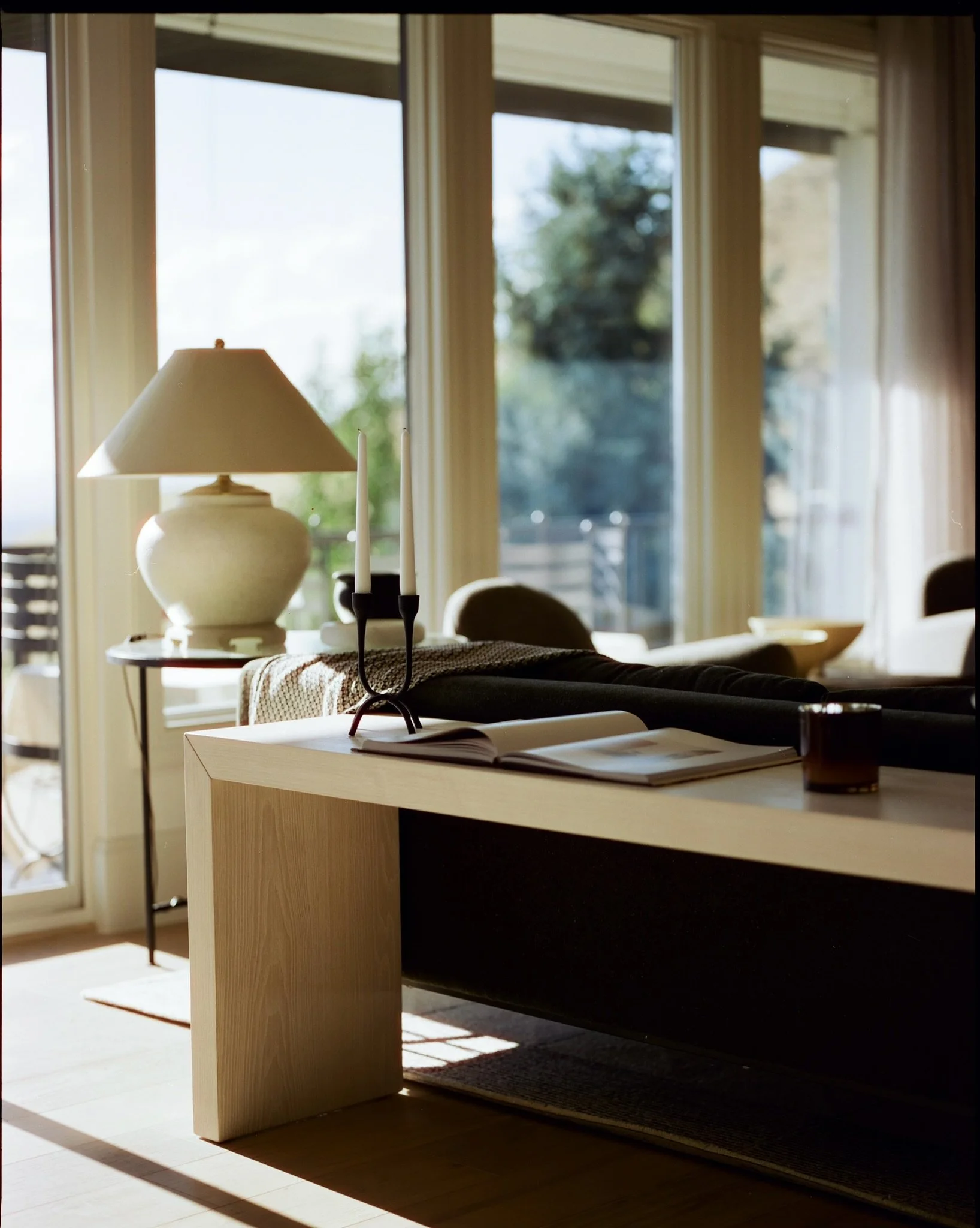 Sunlit modern living room with large windows, a white table lamp, a black candle holder with two candles, an open book, and a glass of drink on a wooden table.