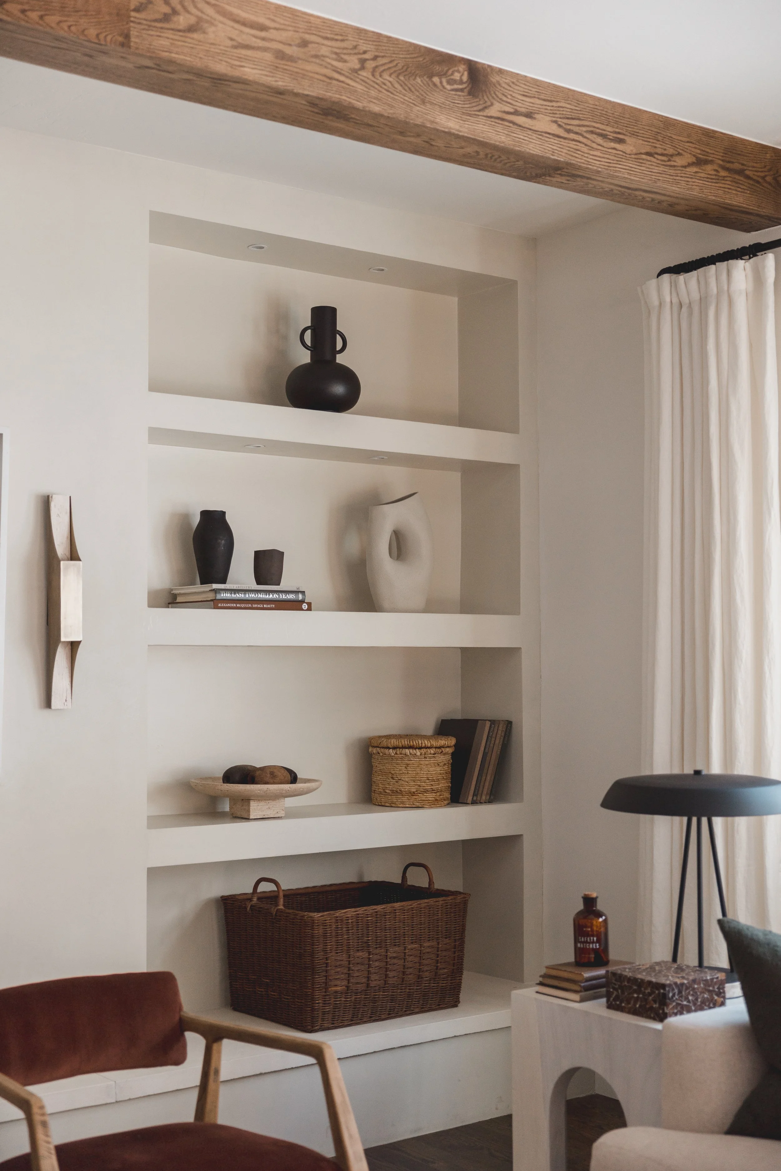 Built-in white wall shelves decorated with black, white, and natural-colored vases, a bowl, books, a wicker basket, and a small sculpture, with a window with cream curtains and a black floor lamp beside.