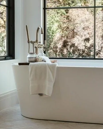 A white freestanding bathtub by a large window with a towel hanging over its side, with trees visible outside.