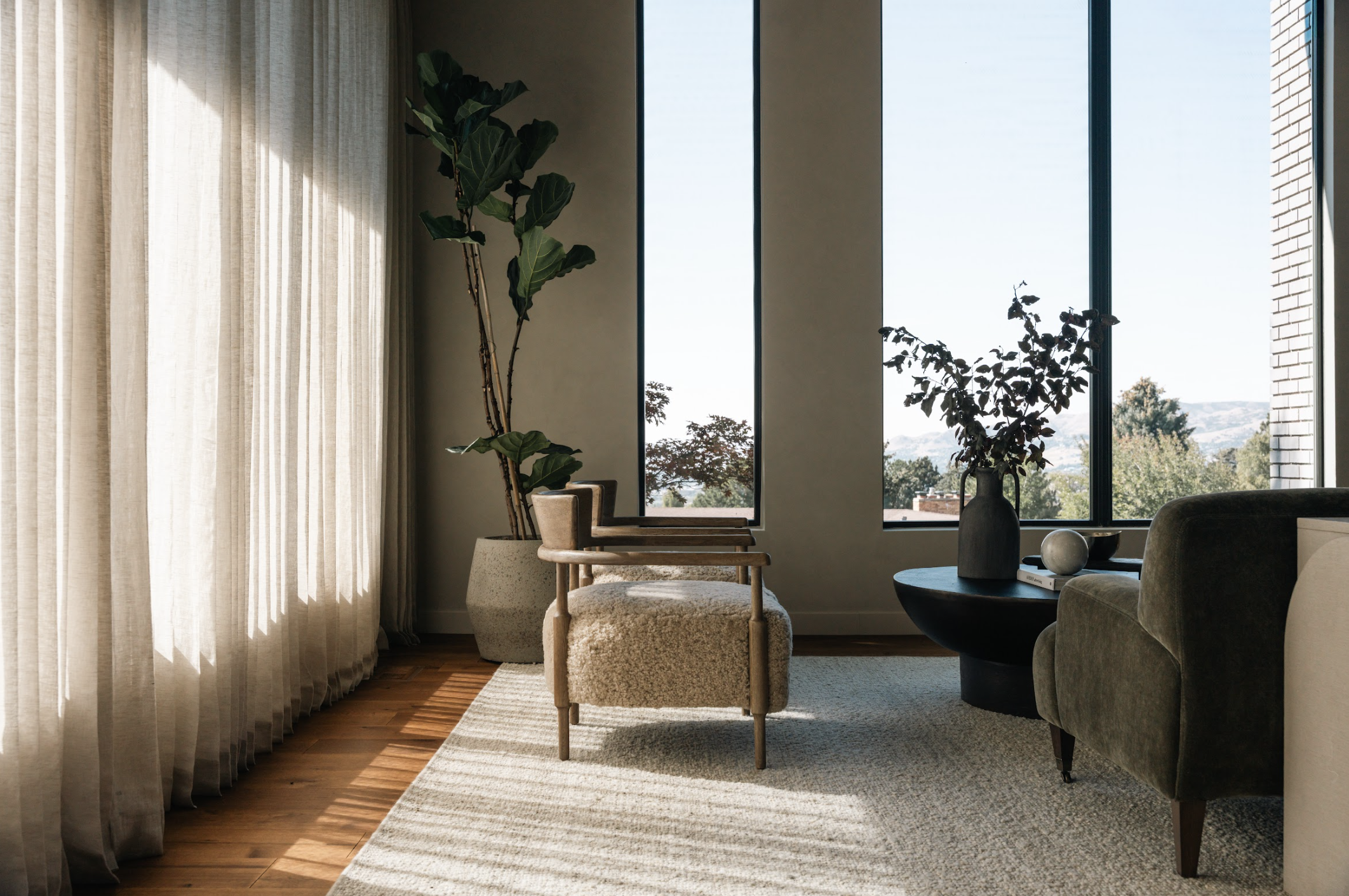 Living room with large windows, beige curtains, a tall potted plant, an armchair with a plush beige cushion, a dark round coffee table with a vase and decorative objects, a green velvet chair, and hardwood floors with a light area rug.