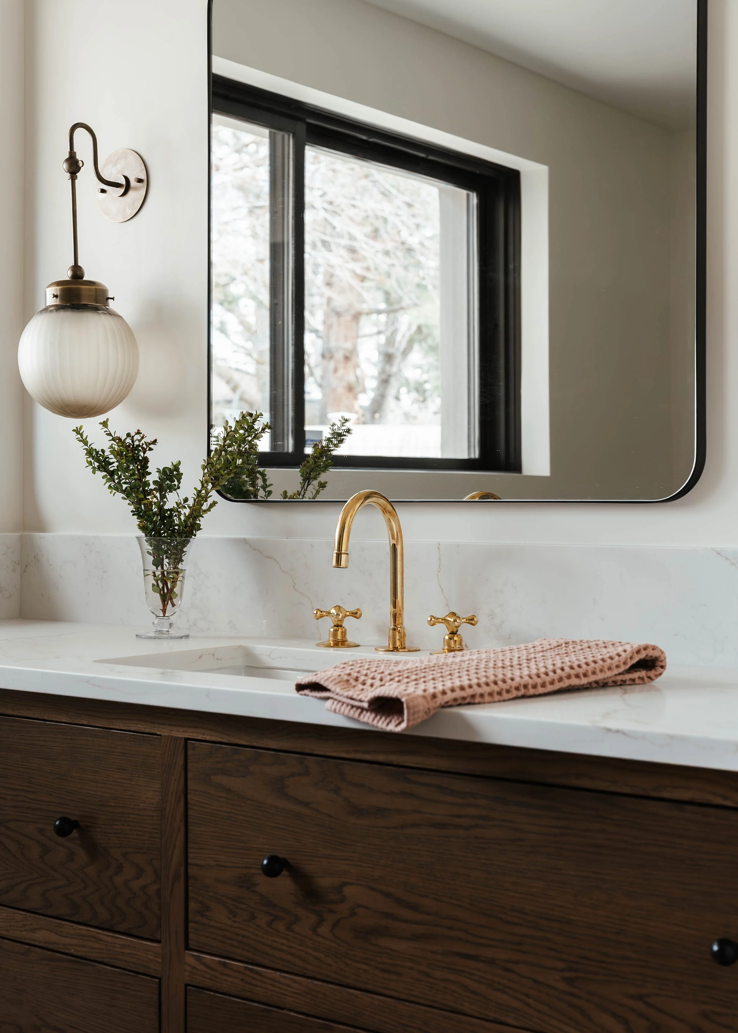 Bathroom sink with a marble countertop, gold faucet, pink hand towel, a vase with green foliage, a mirror, and a wall-mounted light fixture.