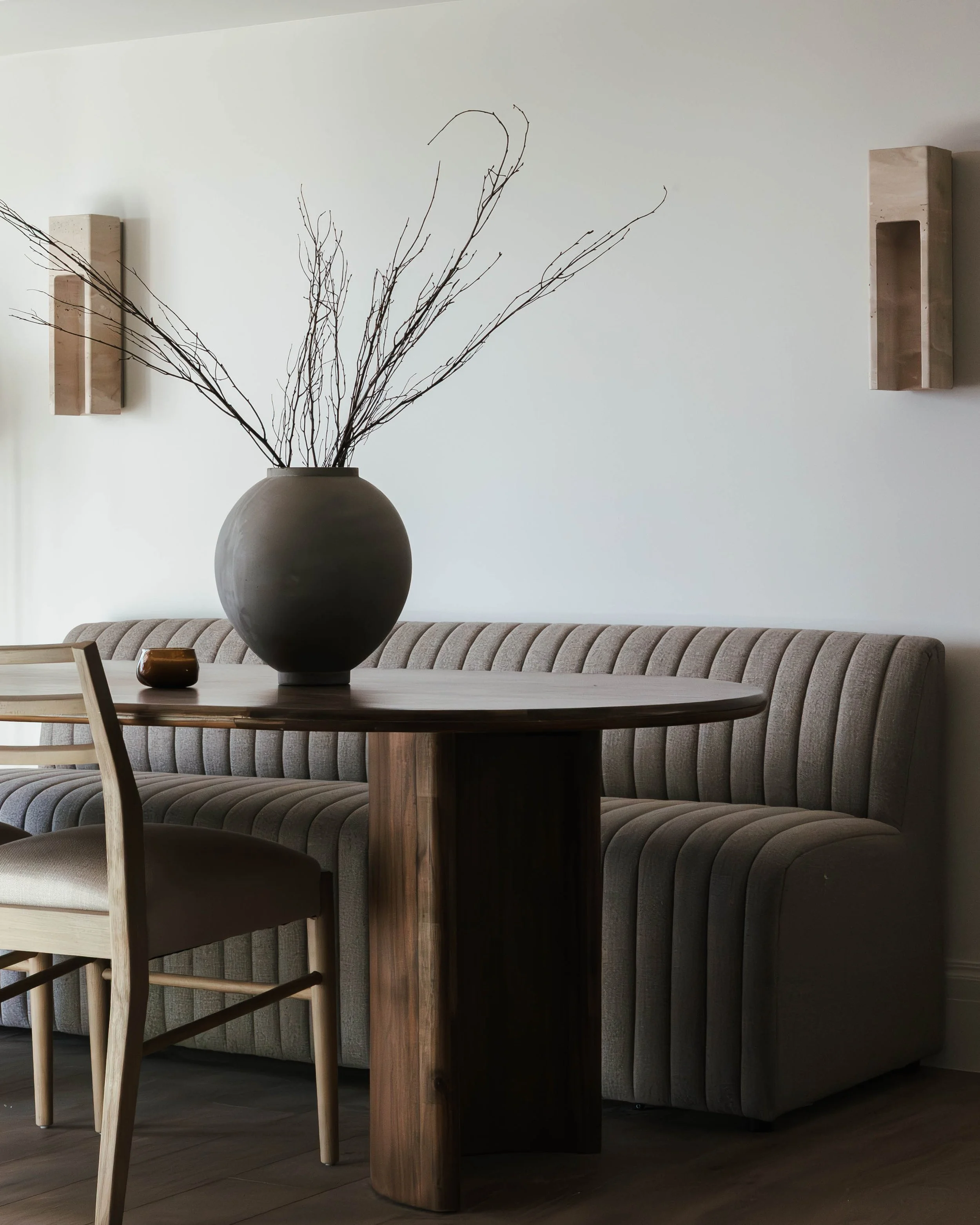 Interior of a room with a round wooden table, a large dark ceramic vase with bare branches, a beige upholstered bench, and two minimalist wall-mounted shelves.