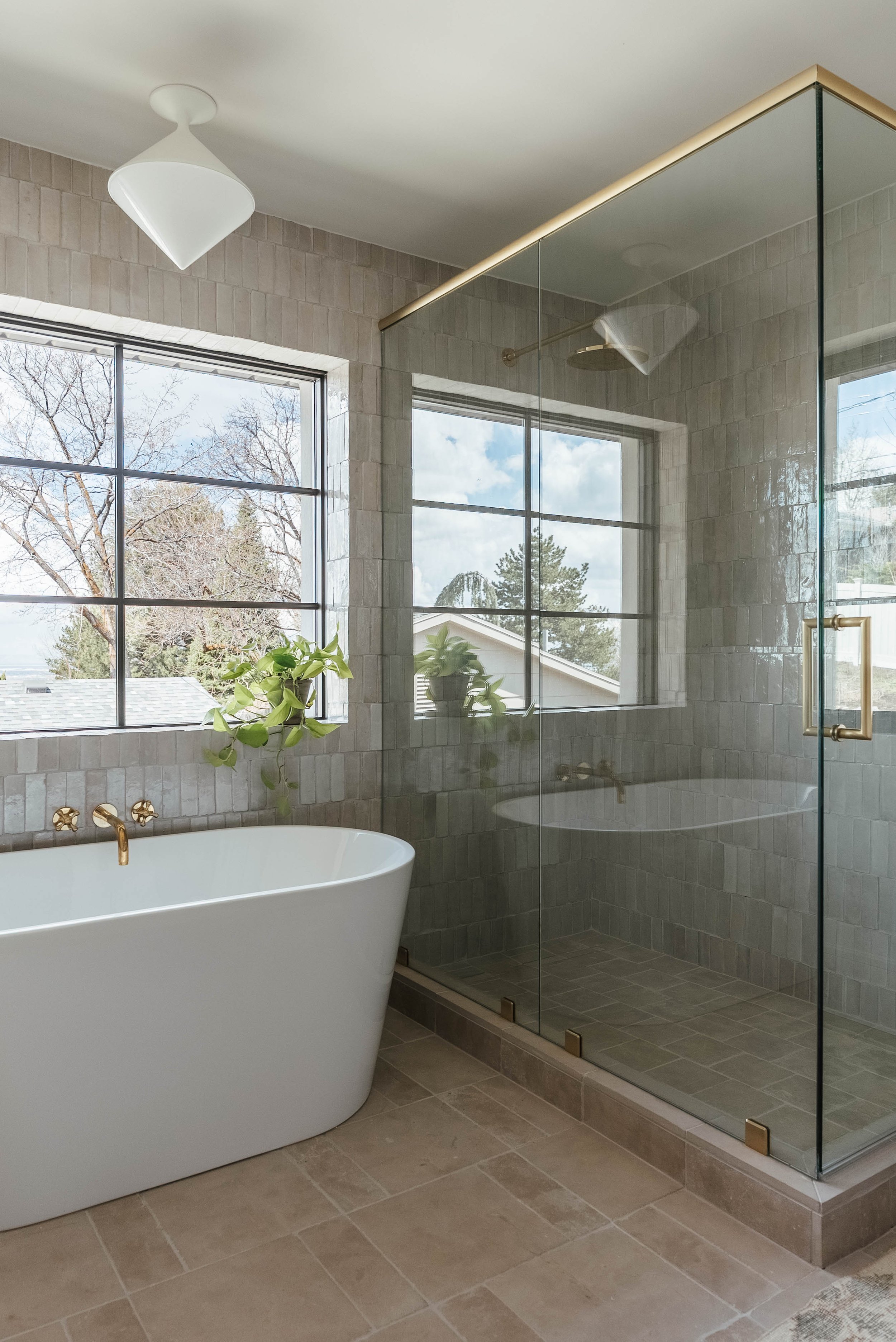 A modern bathroom featuring a white bathtub with gold fixtures and a glass-enclosed shower with a gold handle. There are two large windows with a view of trees and neighboring houses, and potted plants on the windowsill and inside the shower. The bat
