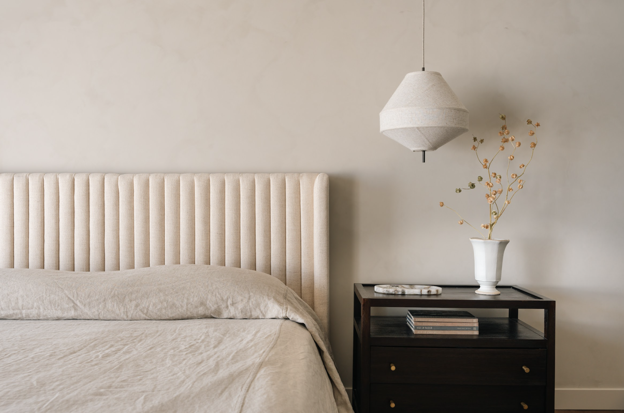 A bedroom with a beige upholstered headboard and bed, a dark wood nightstand with books and a marble tray, a white vase with dried flowers, and a hanging fabric pendant light.
