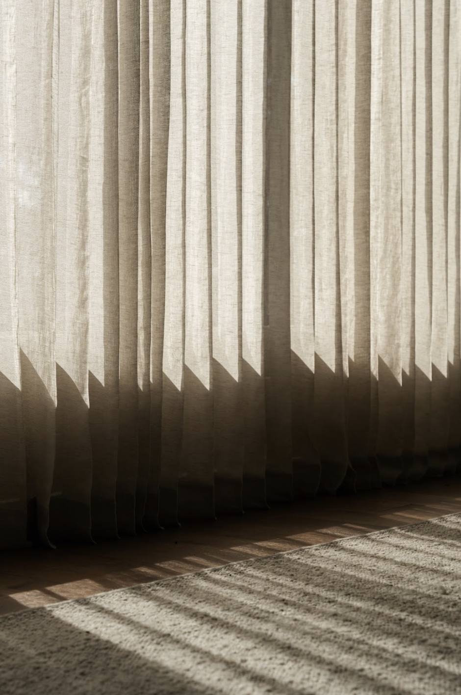 Light beige curtains with sunlight casting shadows on a wooden floor and a textured area rug.