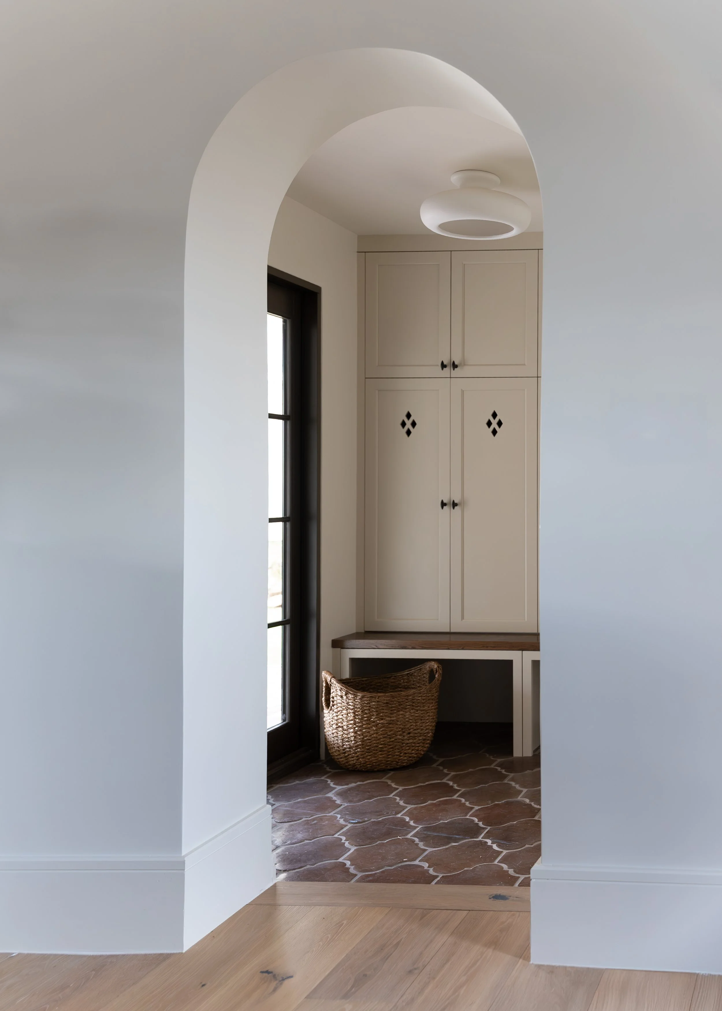 Interior view through an arched doorway, showing a corner with a kitchen cabinet, a window, a ceiling light fixture, a bench, and a basket on a terracotta tiled floor.