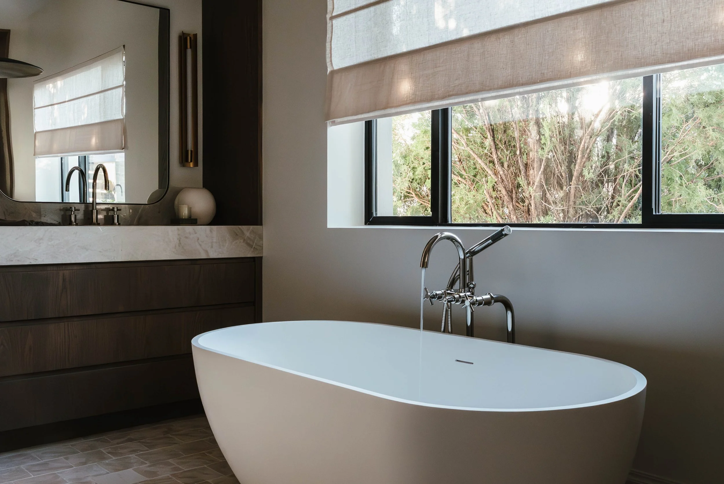 Modern bathroom with freestanding bathtub, large window with beige blinds, wooden vanity with marble countertop, and a large mirror.